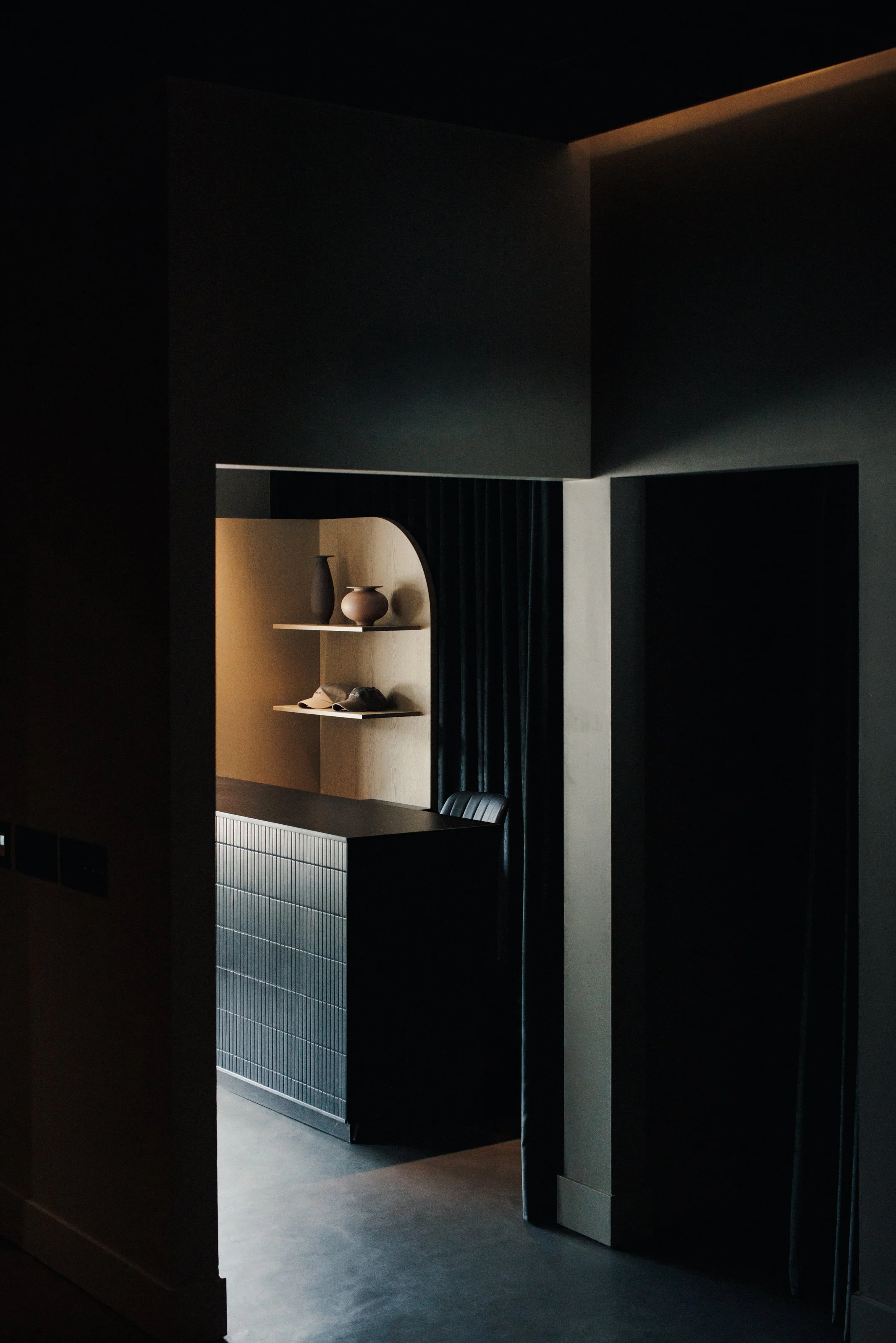 View of reception desk with black tile, black velvet drapery behind and curved pine wall in the background of the photo. Two hats and 2 vases sit on sheves.