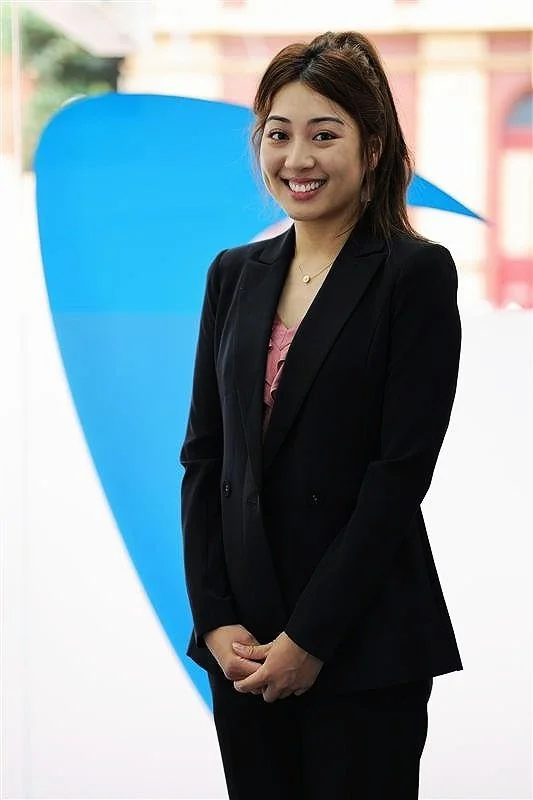 A woman with long dark hair wearing a black blazer and pink top, smiling with her hands clasped in front of her, standing in front of a blue and pink background.