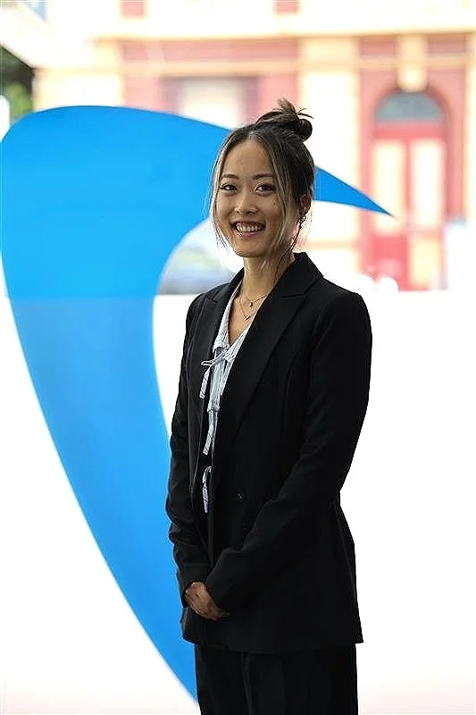 A woman in a black blazer and white blouse is smiling outdoors, standing in front of a large blue curved structure and a building with red accents.