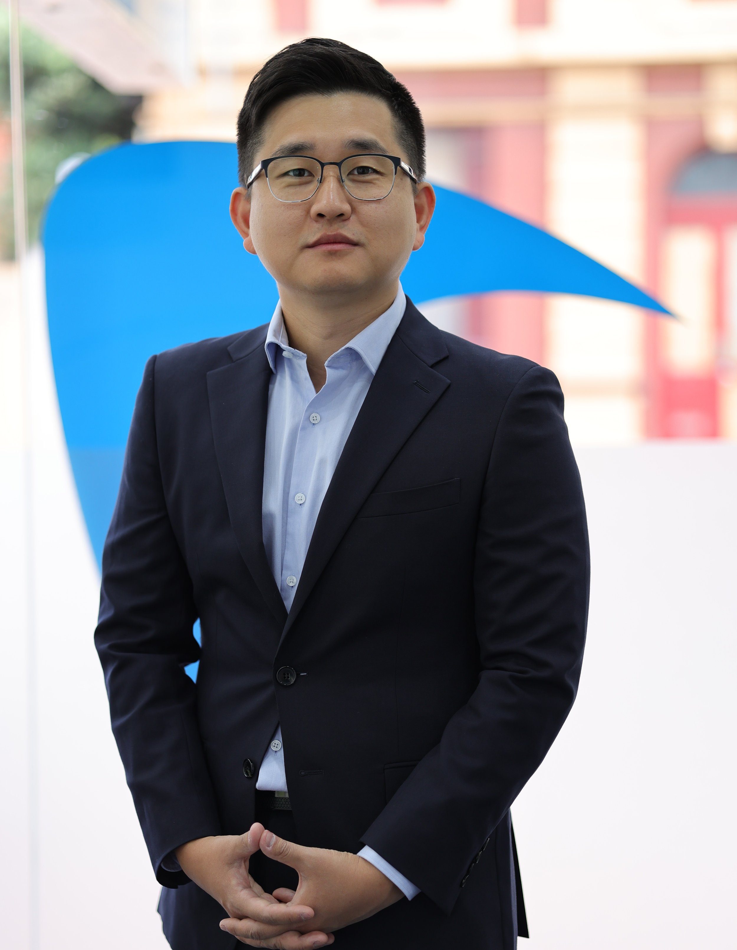 A young Asian man in a dark suit and glasses standing outdoors with a blue abstract sculpture behind him.