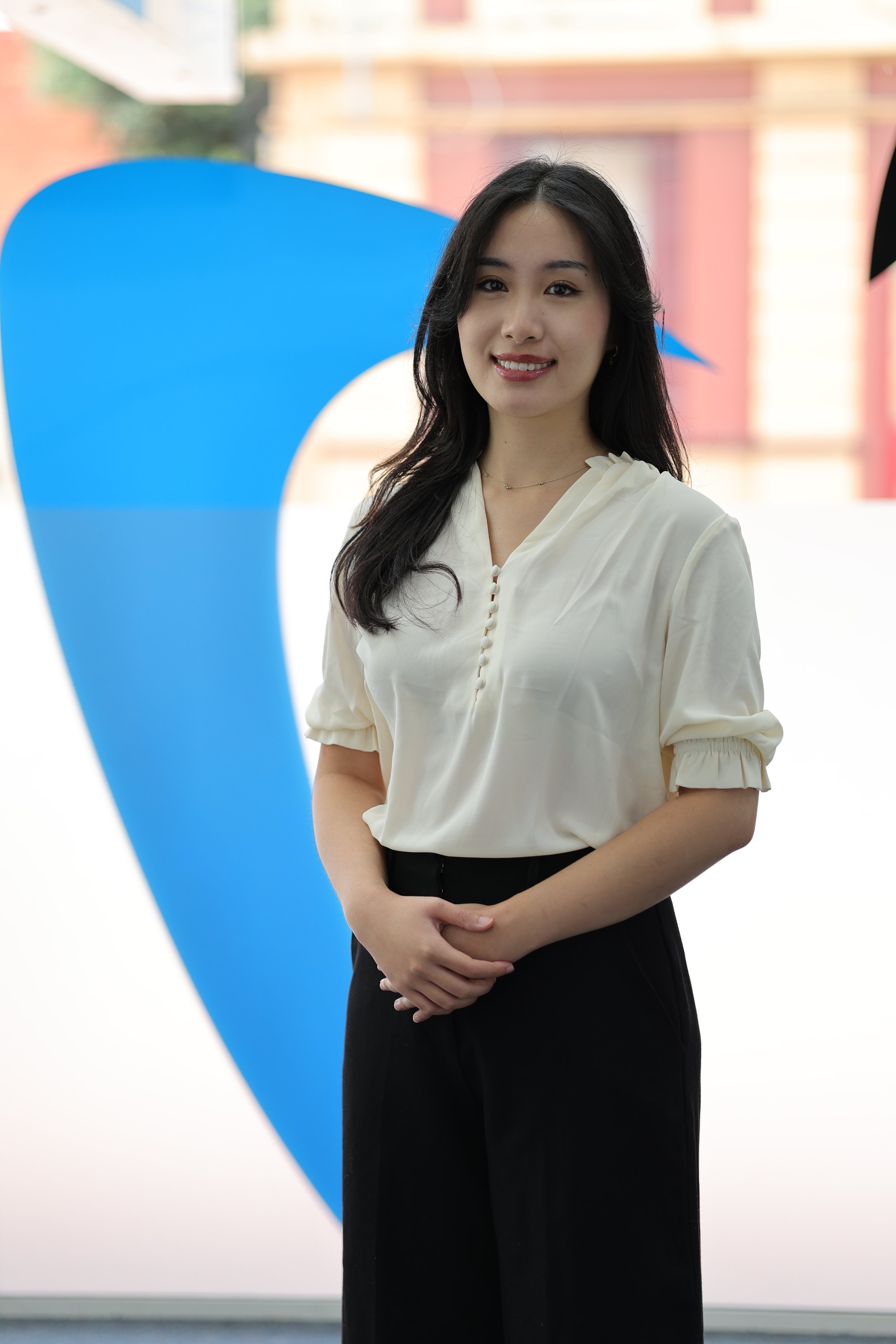 A young woman with long black hair smiling at the camera, standing in front of a large blue abstract shape.