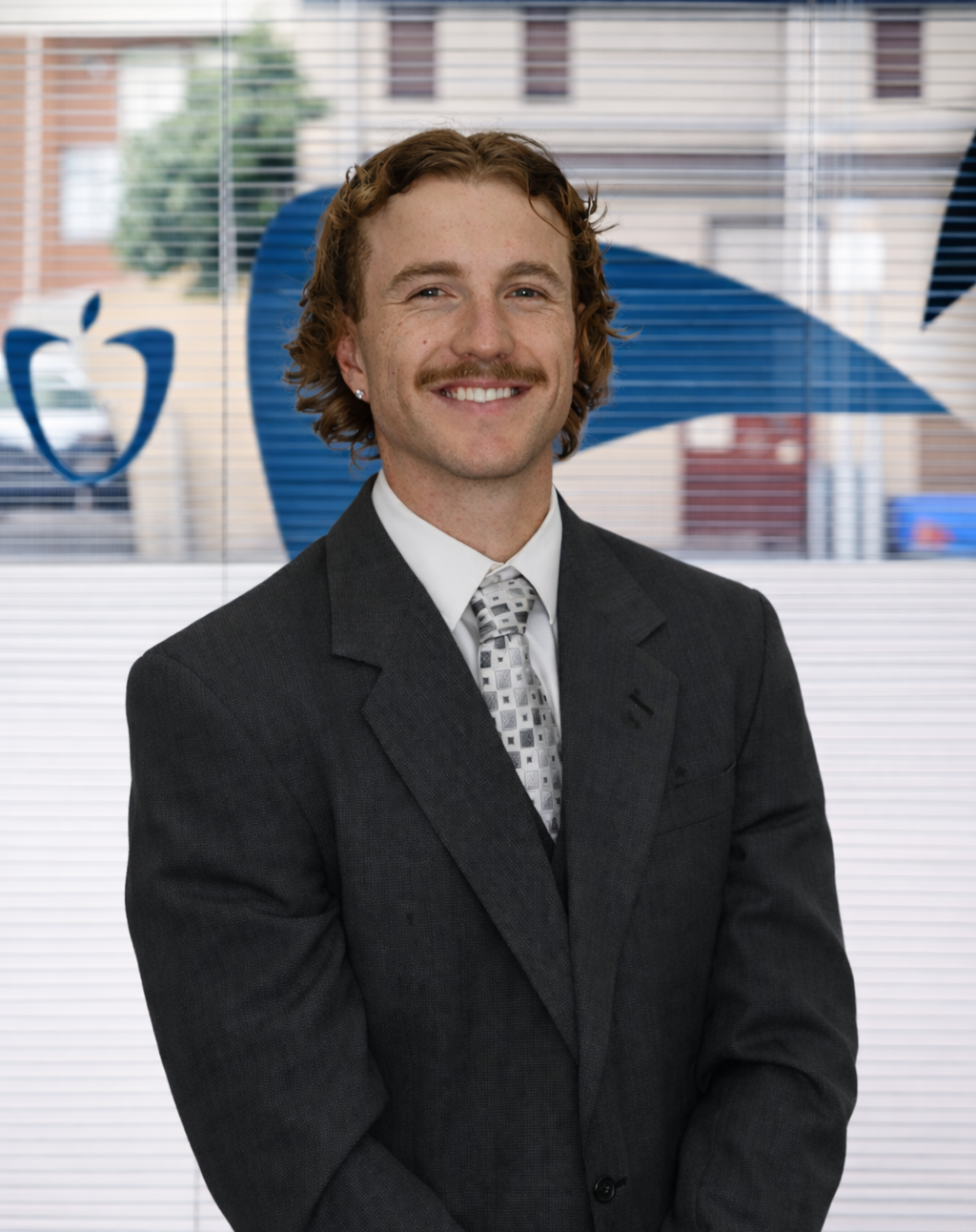 A man with curly red hair, a mustache, wearing a dark suit and a patterned tie, smiling in an office with a large window and a blue company logo in the background.