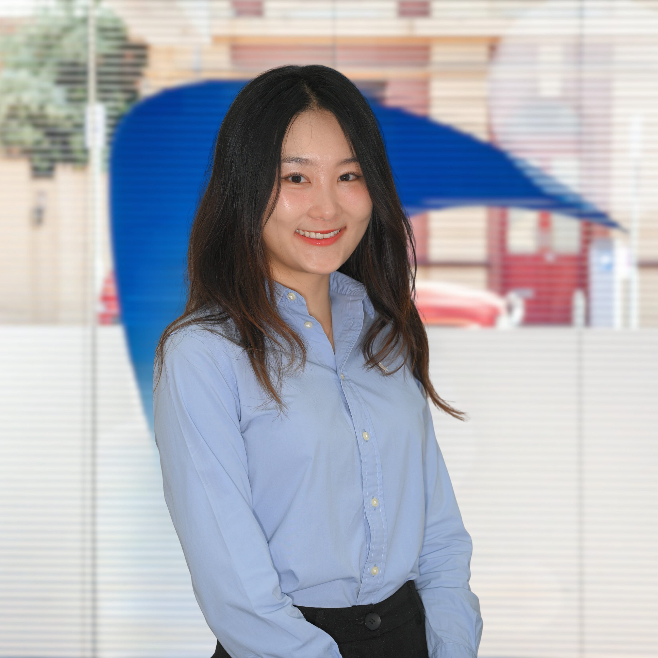 A young woman with long dark hair, wearing a light blue button-up shirt, stands in front of a blurred background with a blue umbrella visible behind her.