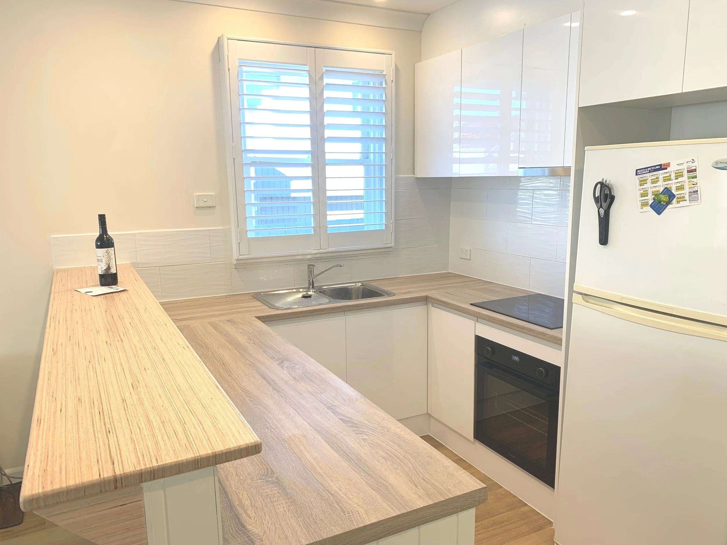 A modern kitchen with light wood countertops, white cupboards, an electric stove, built-in oven, refrigerator, sink under a window with shutters, and a bottle of wine on the counter.
