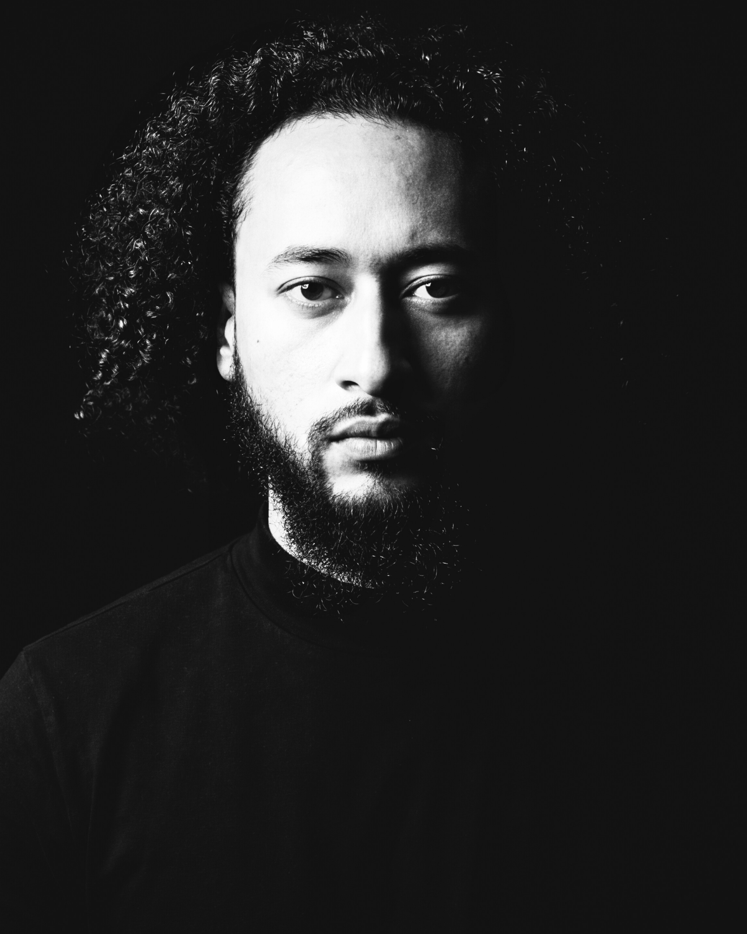 Black and white portrait of a man with curly hair and a beard, wearing a black shirt, against a dark background.