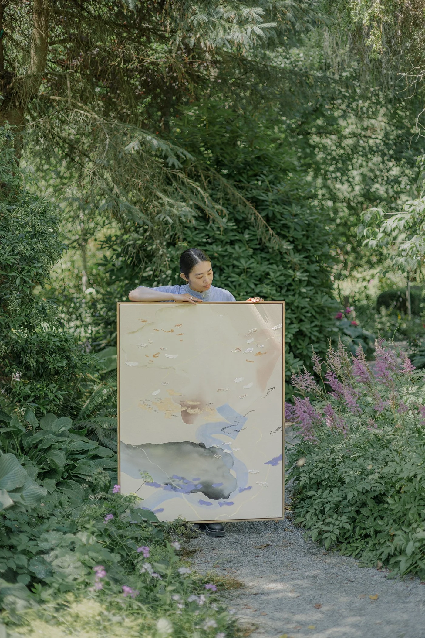 Woman holding a large painting while standing on a garden pathway surrounded by lush green foliage and flowering plants.