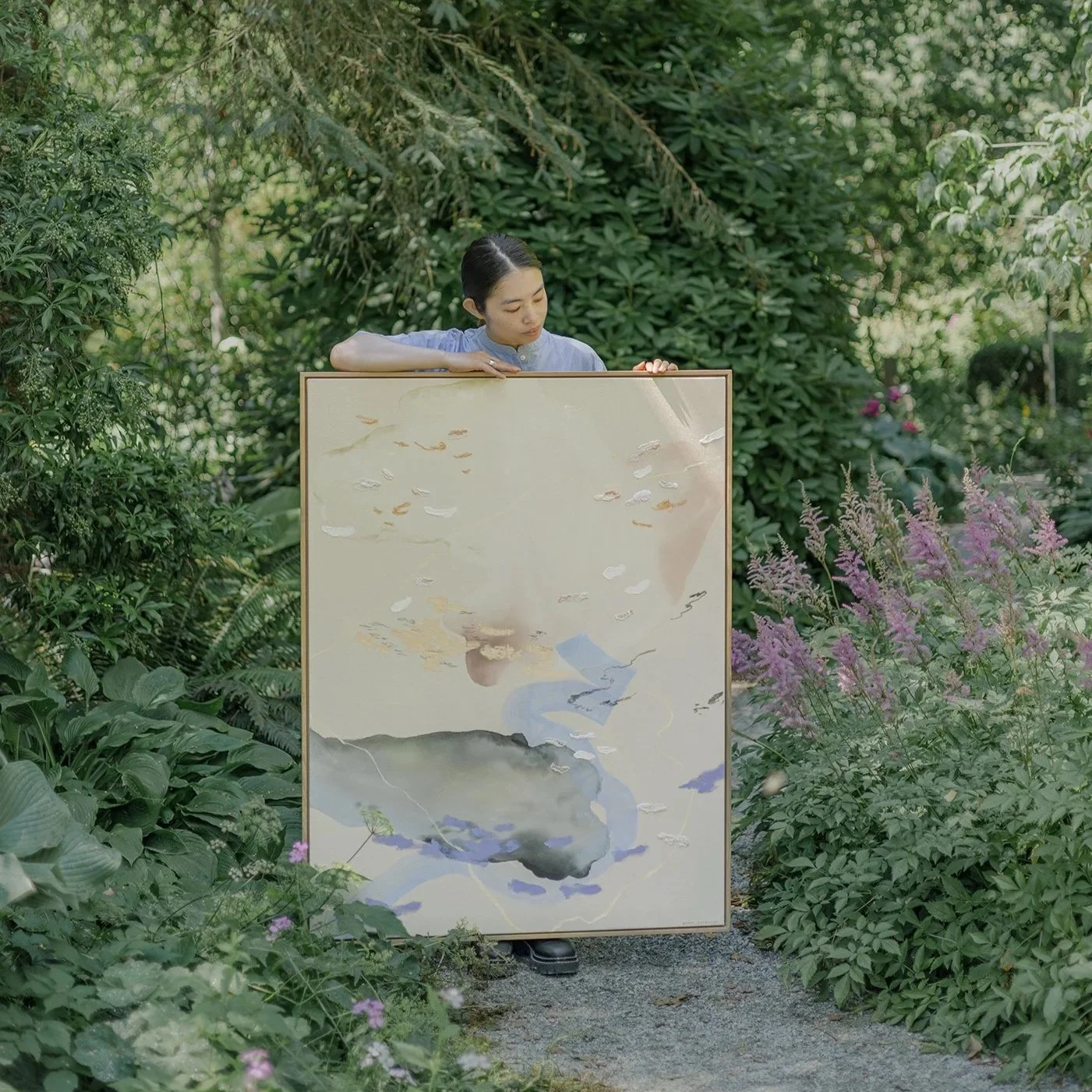 A woman with dark hair in a bun, wearing a light blue shirt, standing in a lush garden surrounded by green plants and purple flowers, holding a large framed painting of a snake and a mountain landscape.