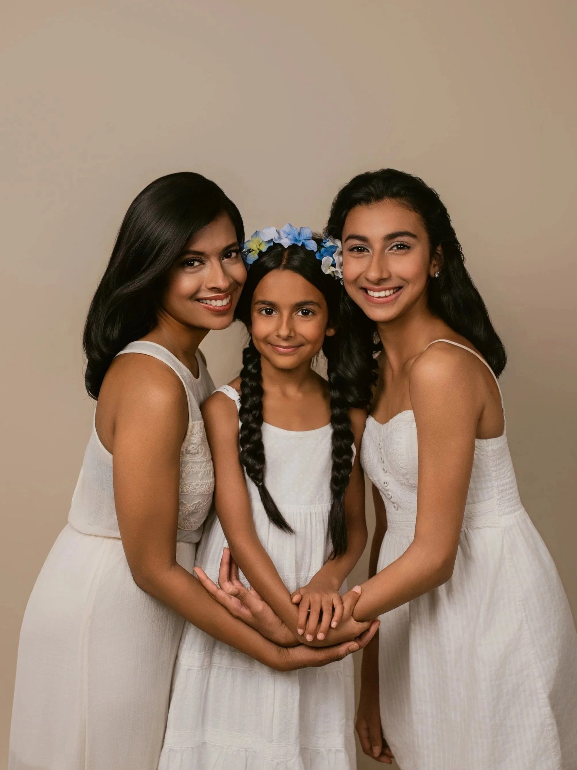 Three women and a girl in white dresses smiling together against a plain background, holding hands.