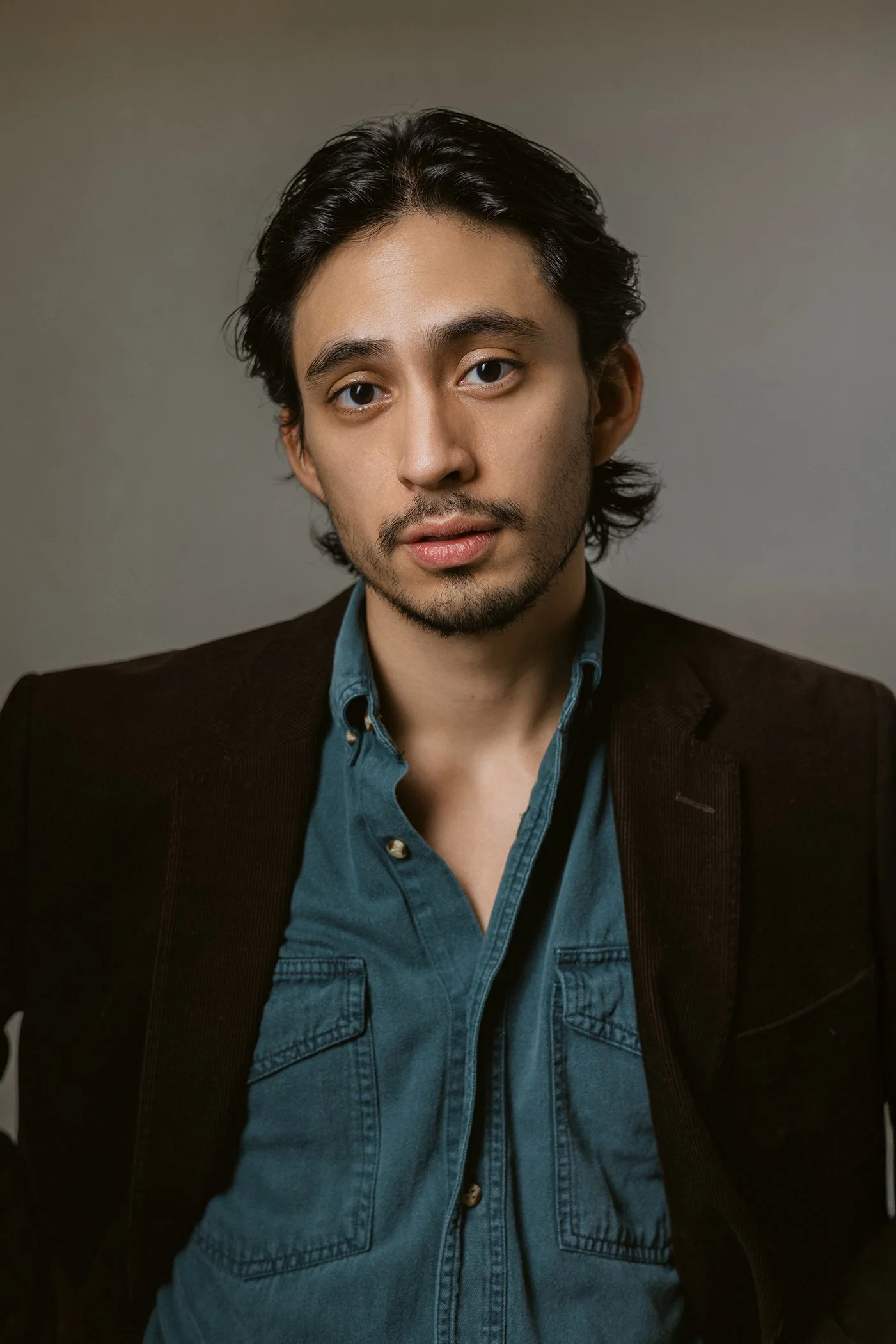 Portrait of a young man with dark hair and facial hair, wearing a denim shirt and a dark blazer, against a plain background.