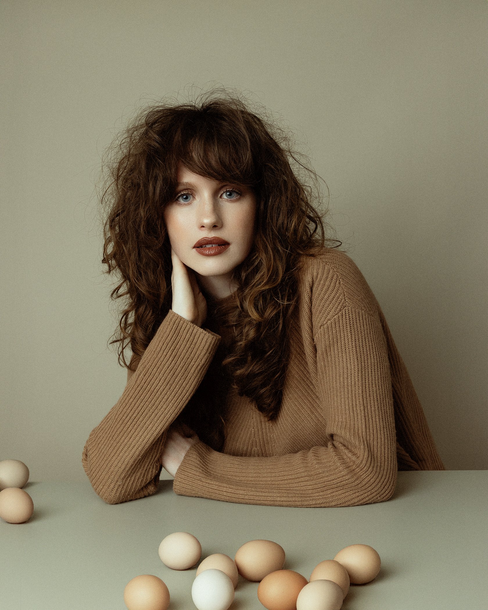 A young woman with curly brown hair and blue eyes resting her chin on her hand, sitting at a table with eggs scattered on it, wearing a tan sweater against a neutral background.