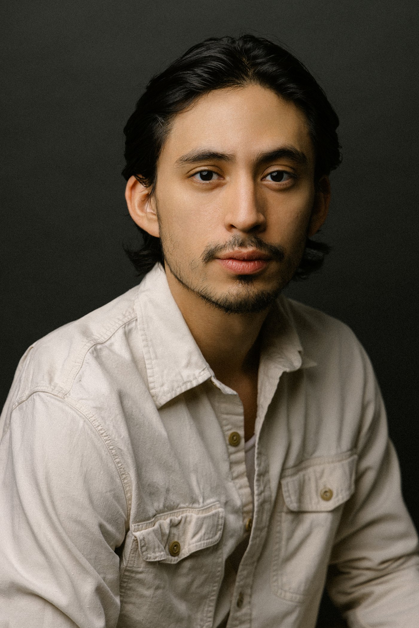 Portrait of a young man with dark hair and facial hair, wearing a light-colored button-up shirt, against a dark background.
