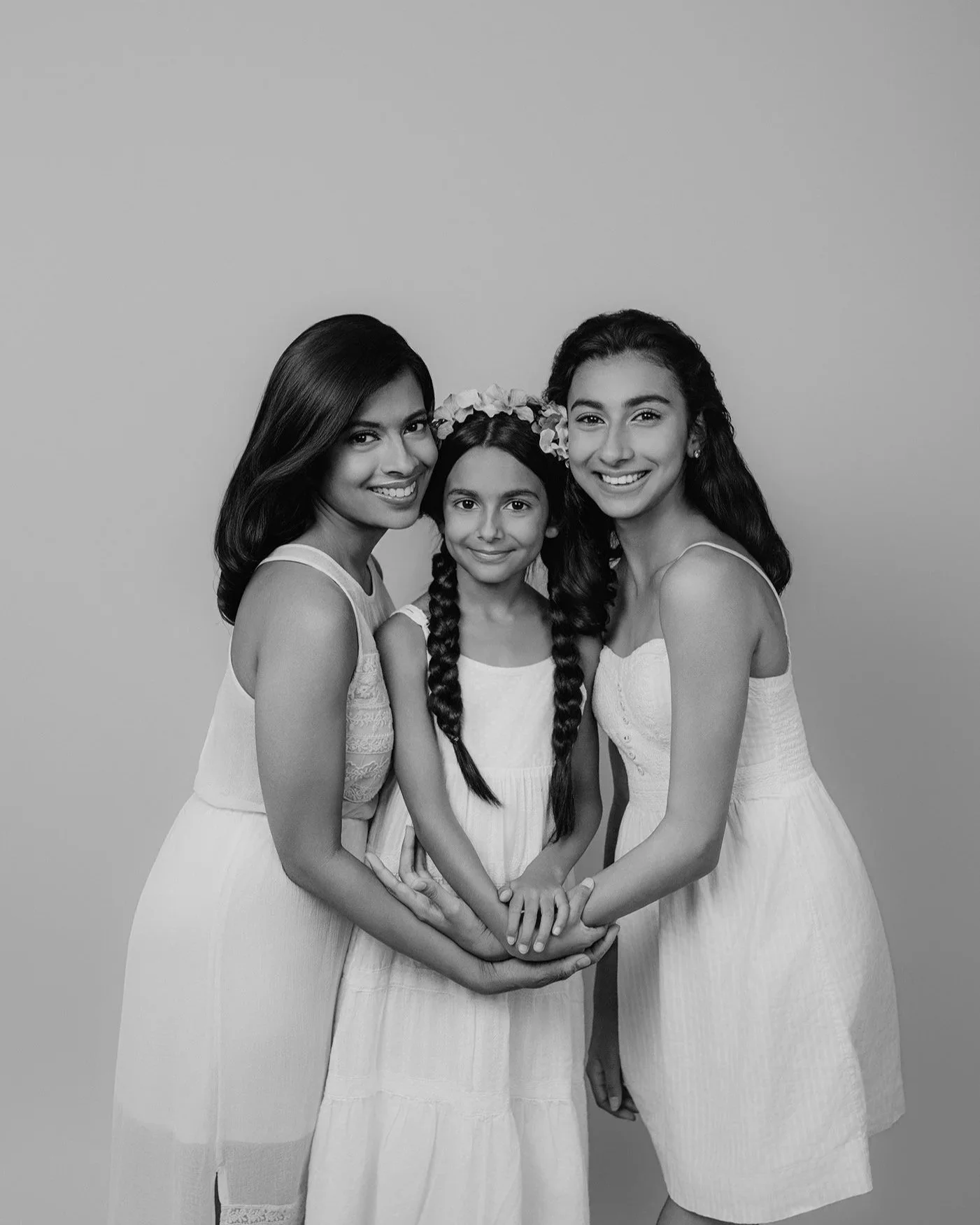 Black and white photo of three smiling girls standing close together, holding hands, with a plain background.