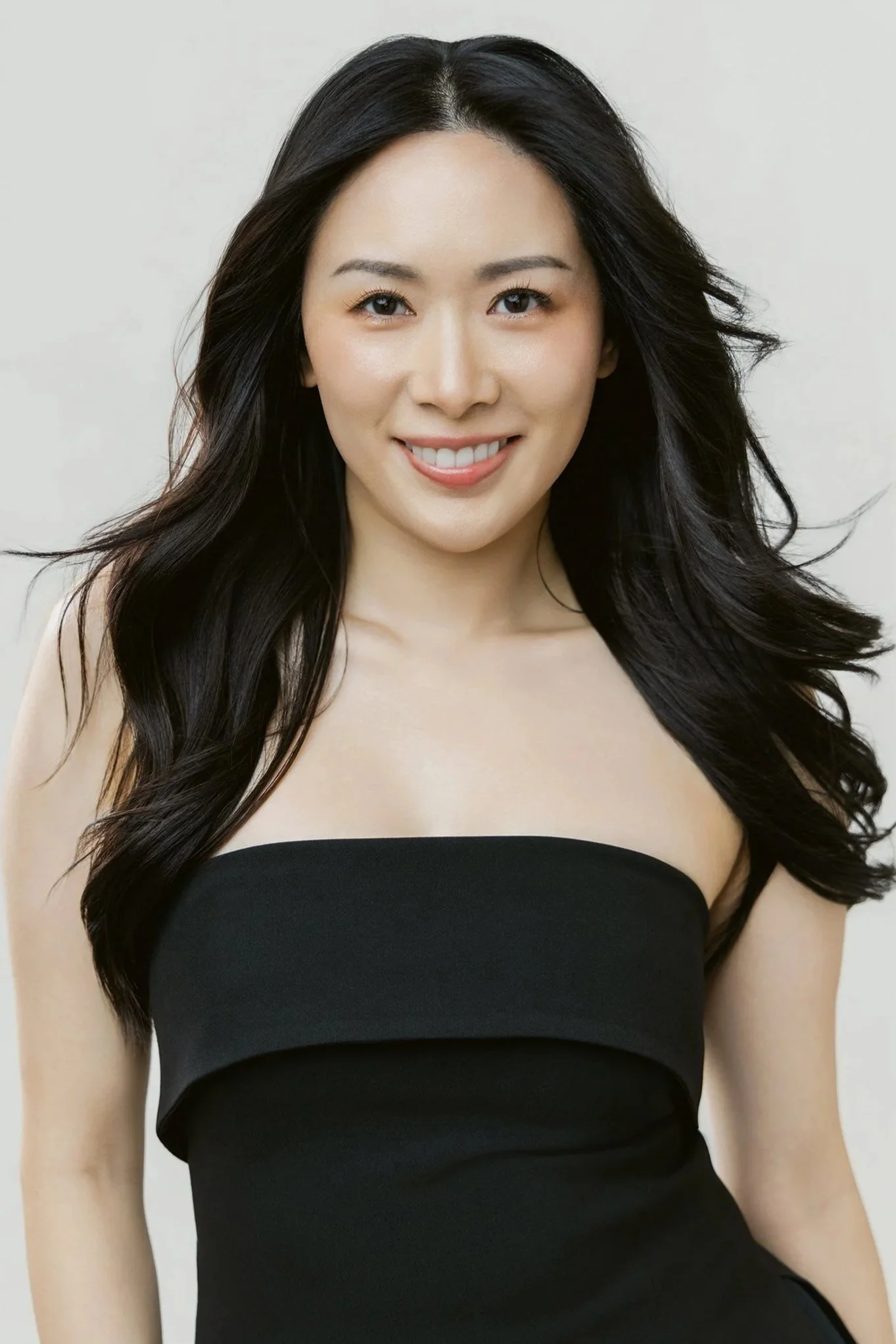 A woman with long dark hair in loose waves, wearing a strapless black dress, smiling at the camera against a plain background.