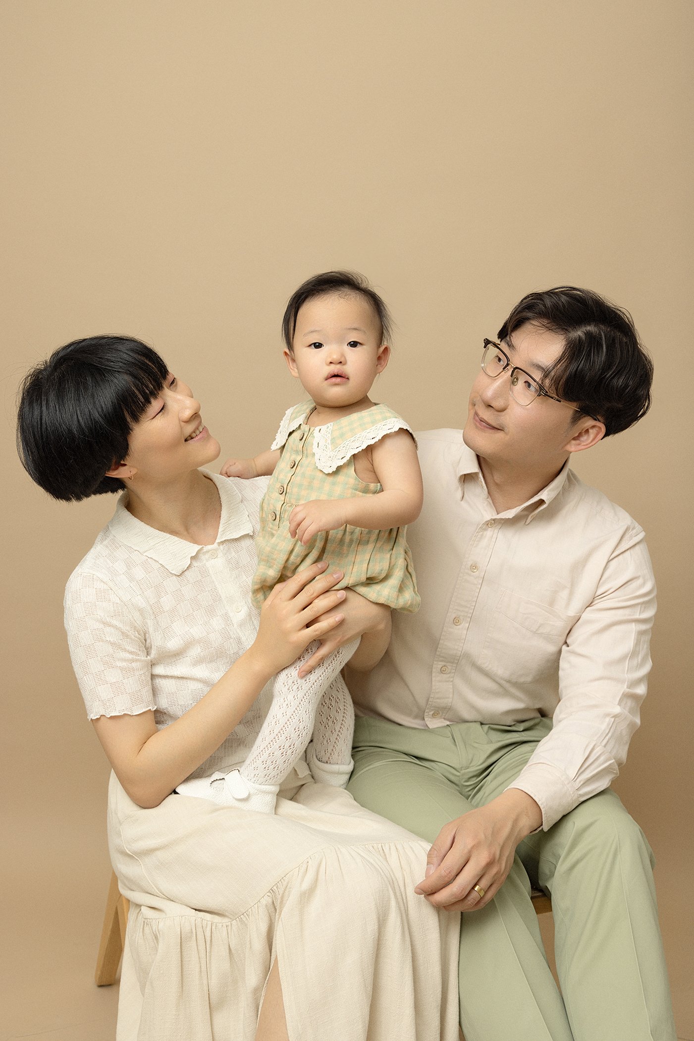 A family of three, a woman, a man, and a young girl, posing against a beige background. The woman is smiling and holding the girl on her lap, while the man is looking at the girl with a gentle expression.