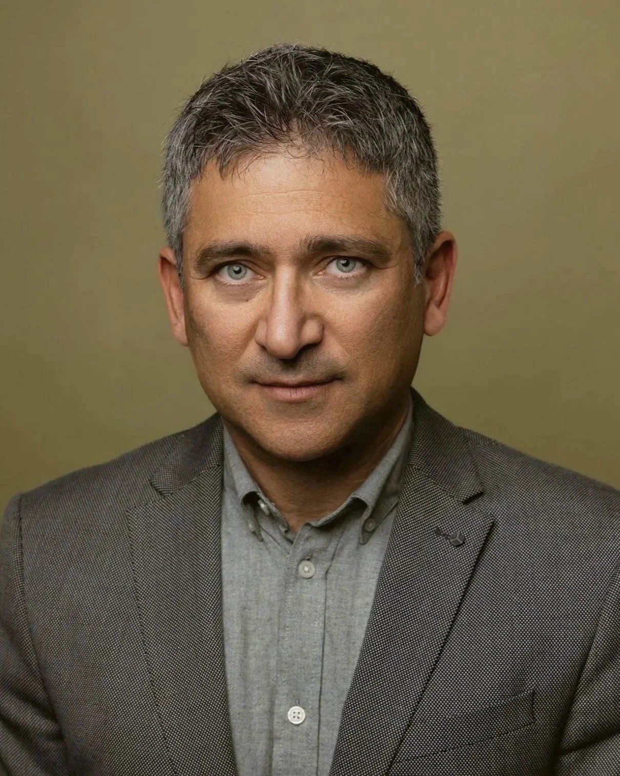 A portrait of a middle-aged man with short, gray hair and light-colored eyes, wearing a gray shirt and a gray blazer, against an olive green background.