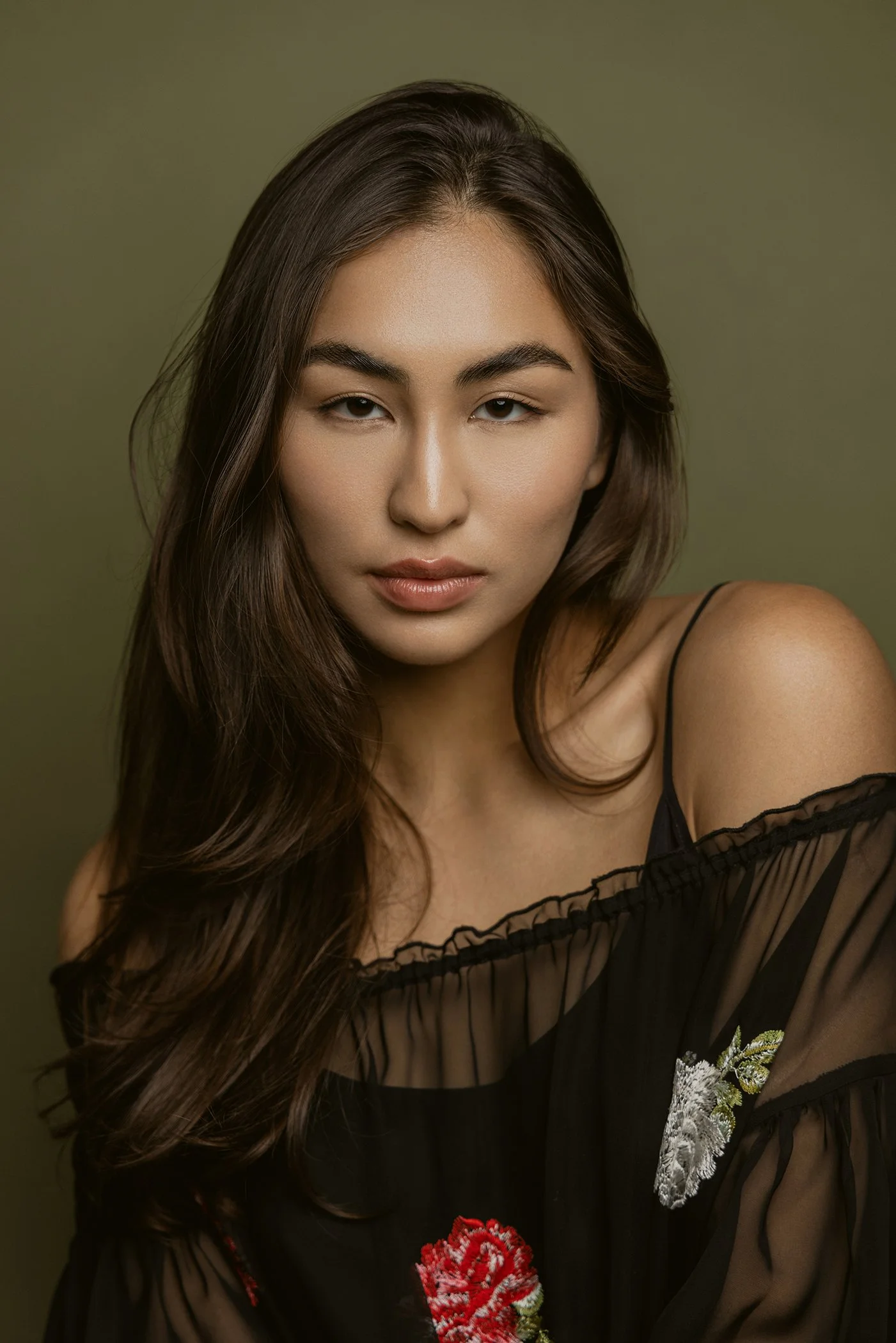 A woman with long, wavy brown hair posing against a plain olive green background, wearing a black sheer top with floral embroidery.