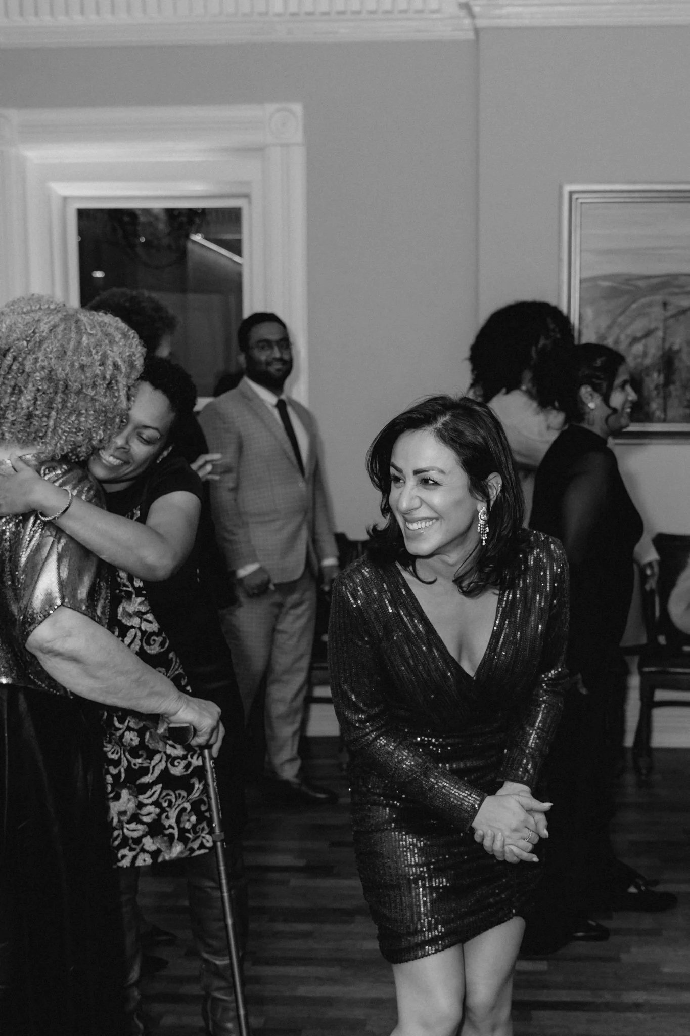 A group of people at a social gathering, including a woman in a black sparkly dress shaking hands with a woman with gray curly hair, other guests are seen smiling and mingling in the background.