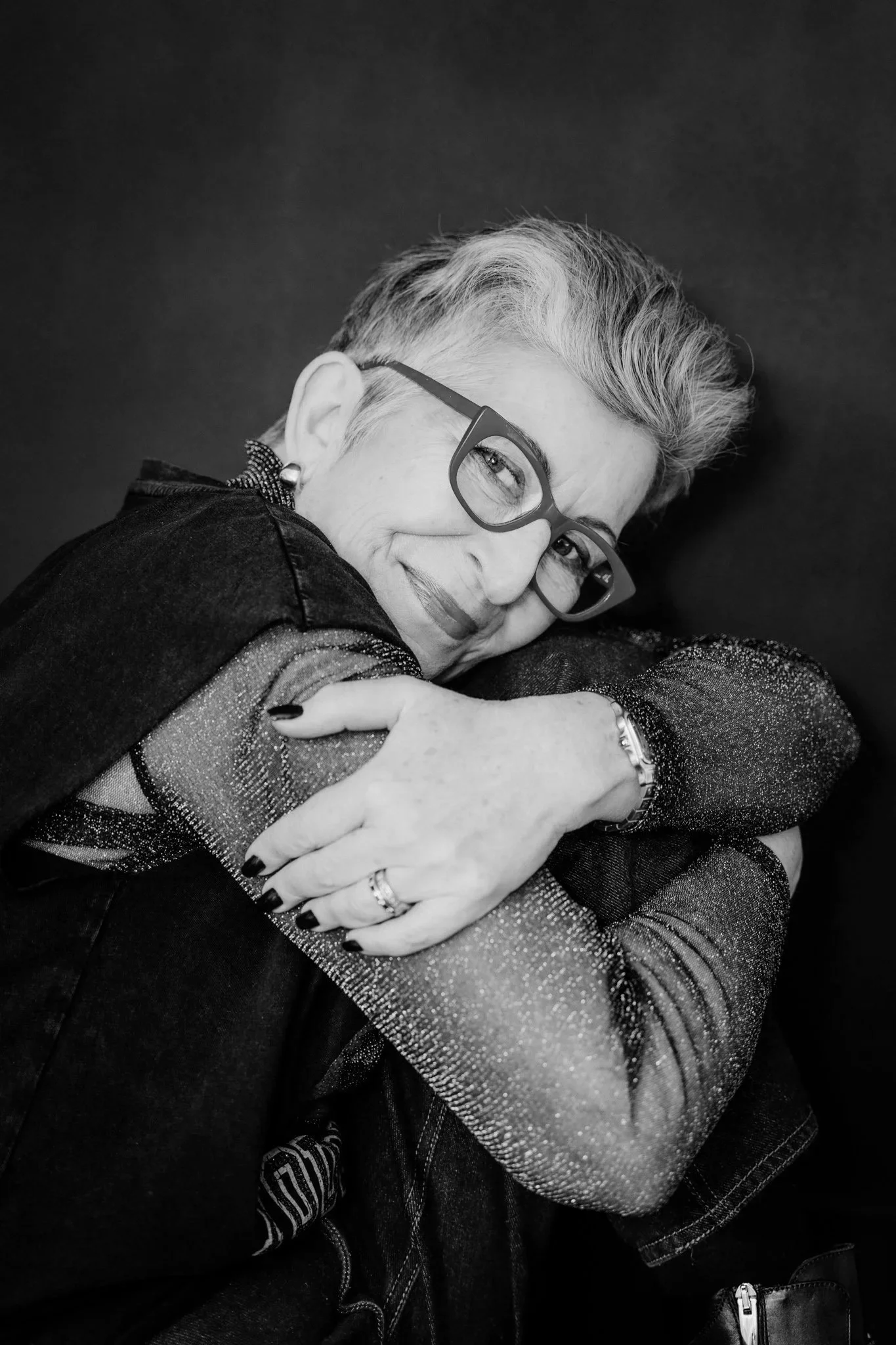 A black and white photo of a woman with short hair and glasses, smiling and hugging someone.