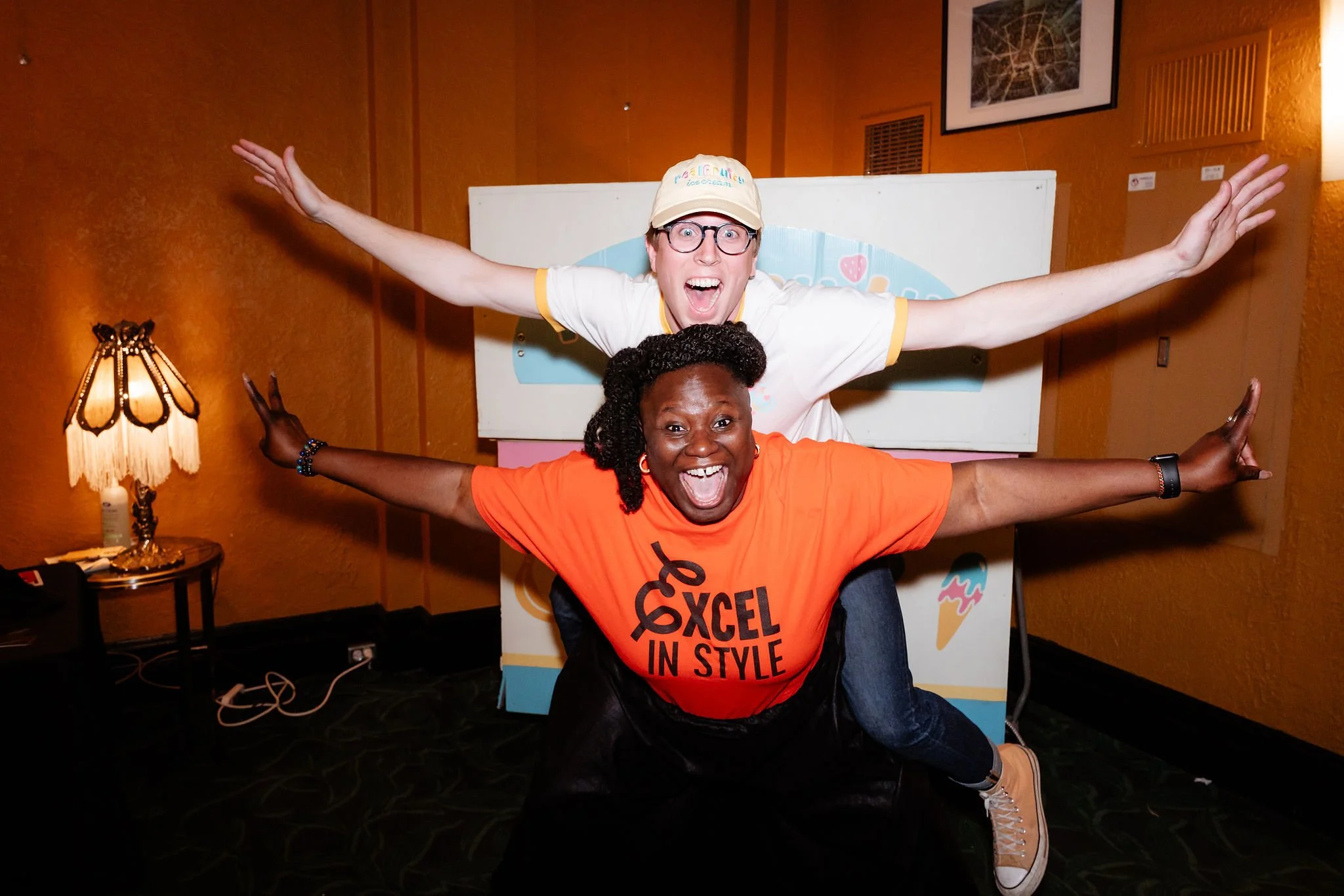 Two excited people in a fun pose, one standing behind the other, both with arms outstretched, inside a room with warm lighting and wooden paneling on the walls.