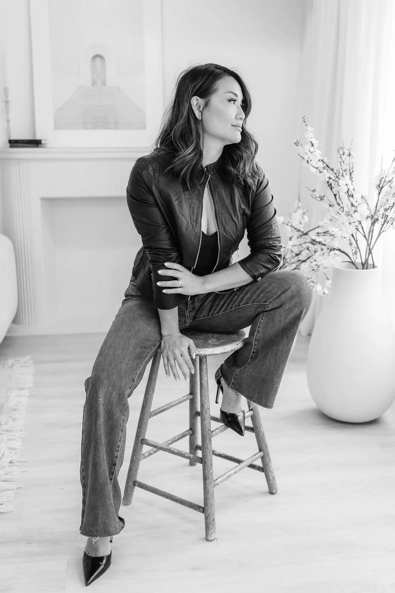 A woman with wavy hair sitting on a wooden stool in a room with a light-colored wooden floor, wearing a leather jacket, jeans, and high heels, with a large potted plant and artwork in the background.