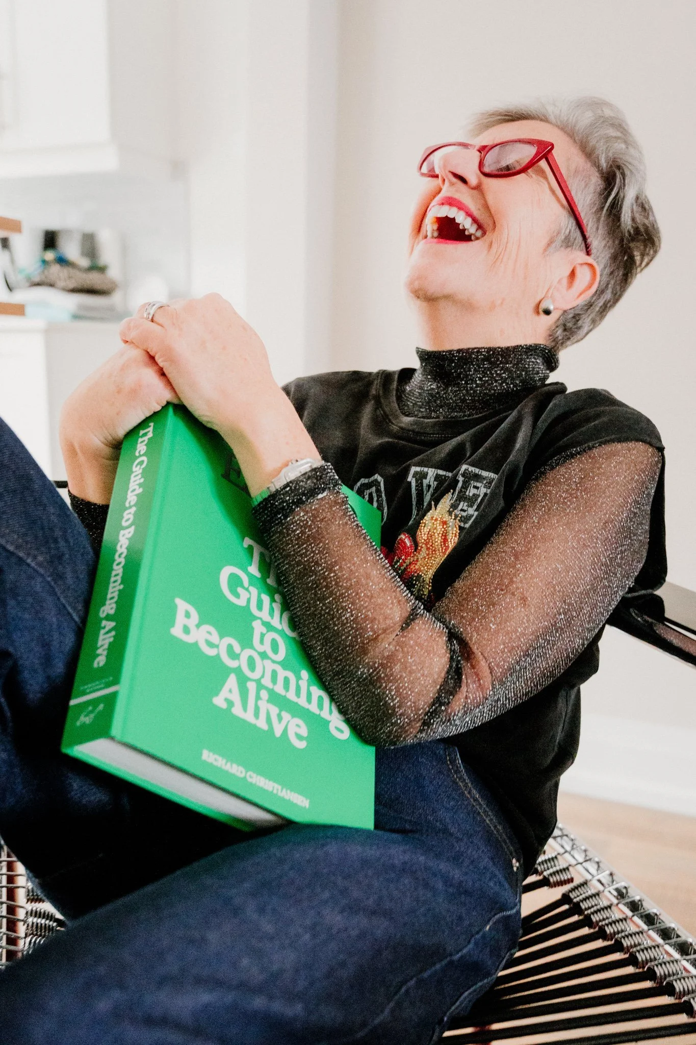 Older woman with short gray hair, wearing red glasses and a black sparkly top, sitting on a chair, smiling and laughing while holding a green book.