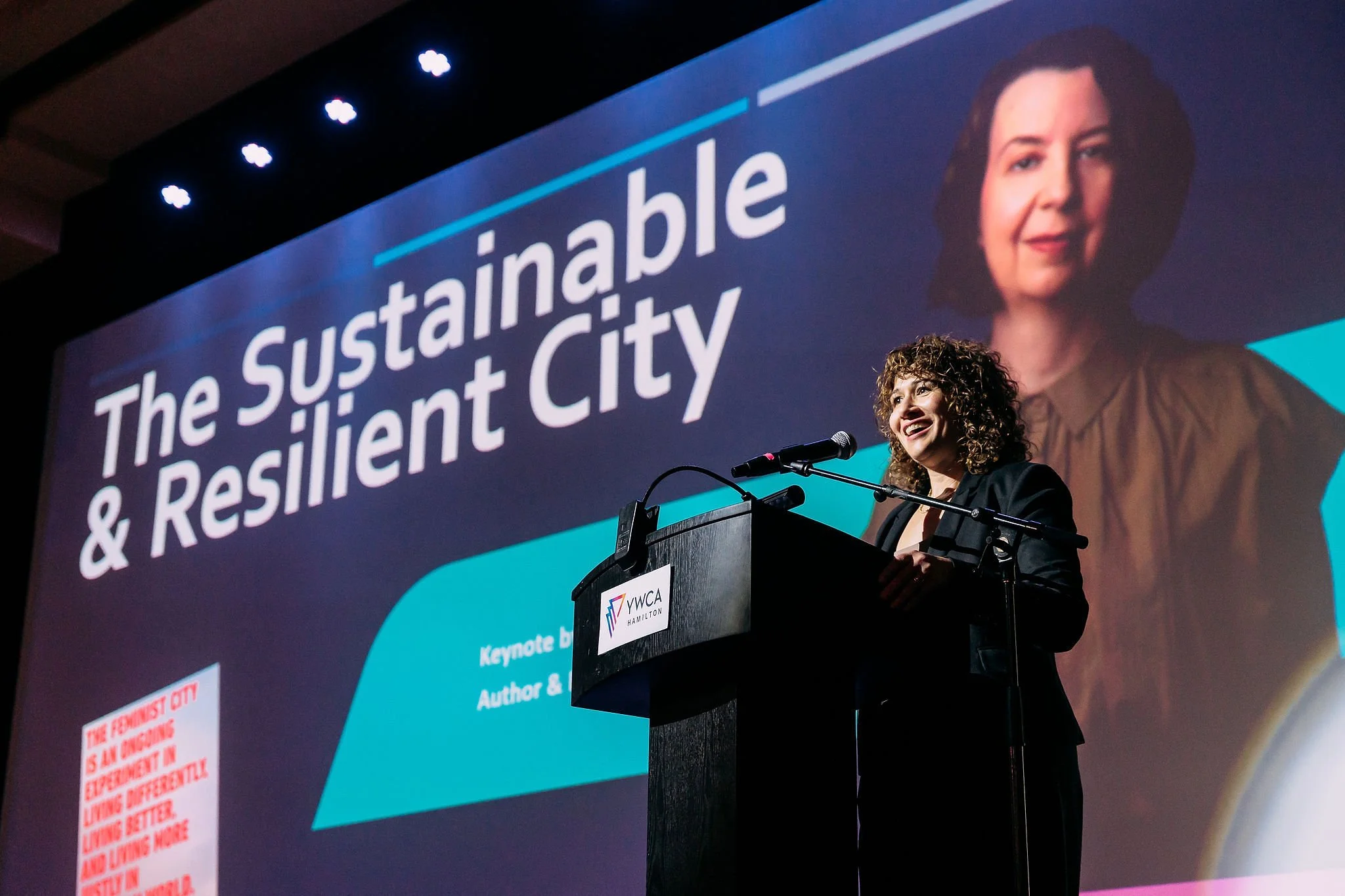 Woman with curly hair speaking at a podium with a microphone, in front of a large screen displaying the words 'The Sustainable & Resilient City' and an image of a woman with shoulder-length dark hair. The podium has a logo for YWCA Hamilton.