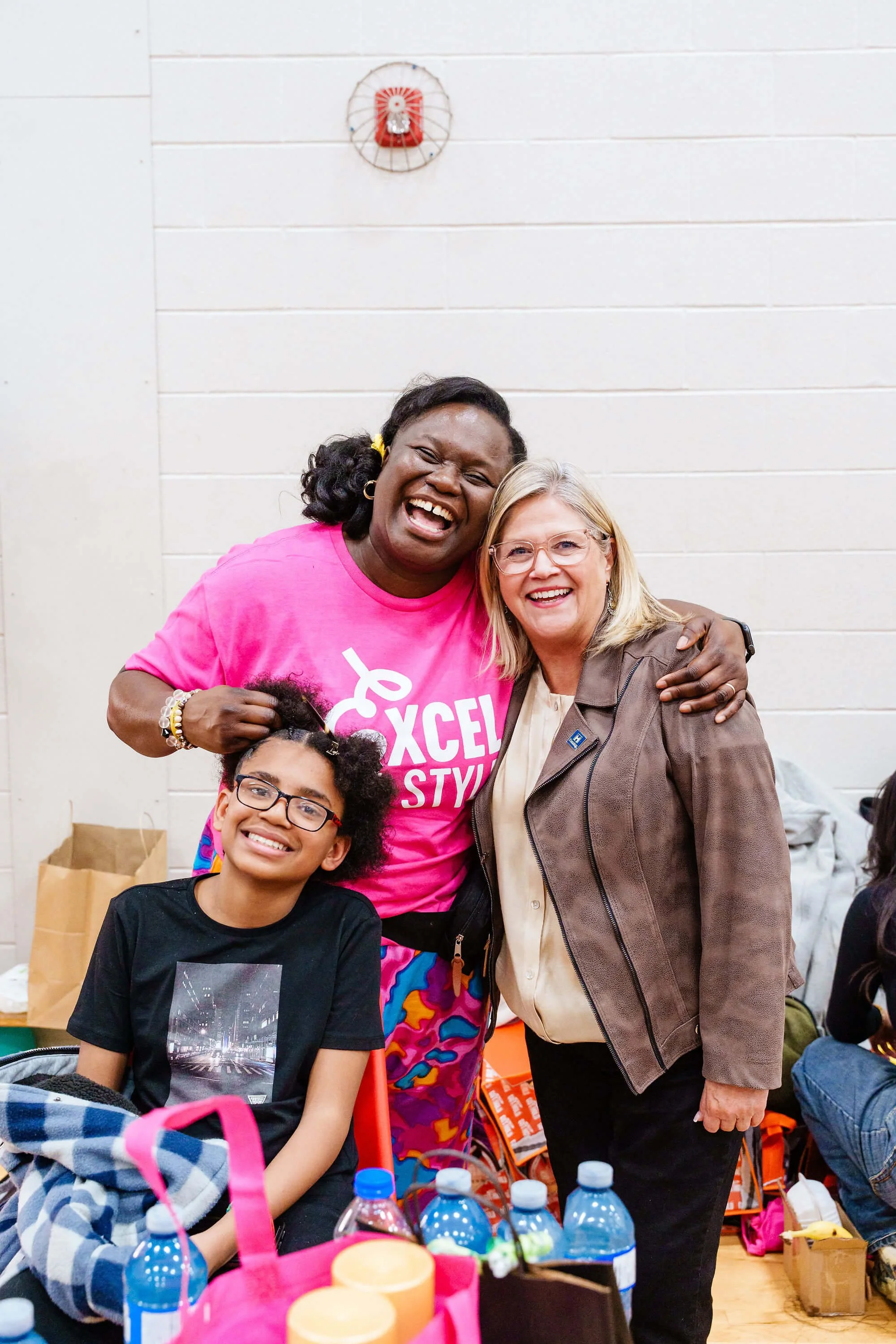 Group of four women, smiling and embracing in an indoor setting. One woman is wearing a bright pink t-shirt with