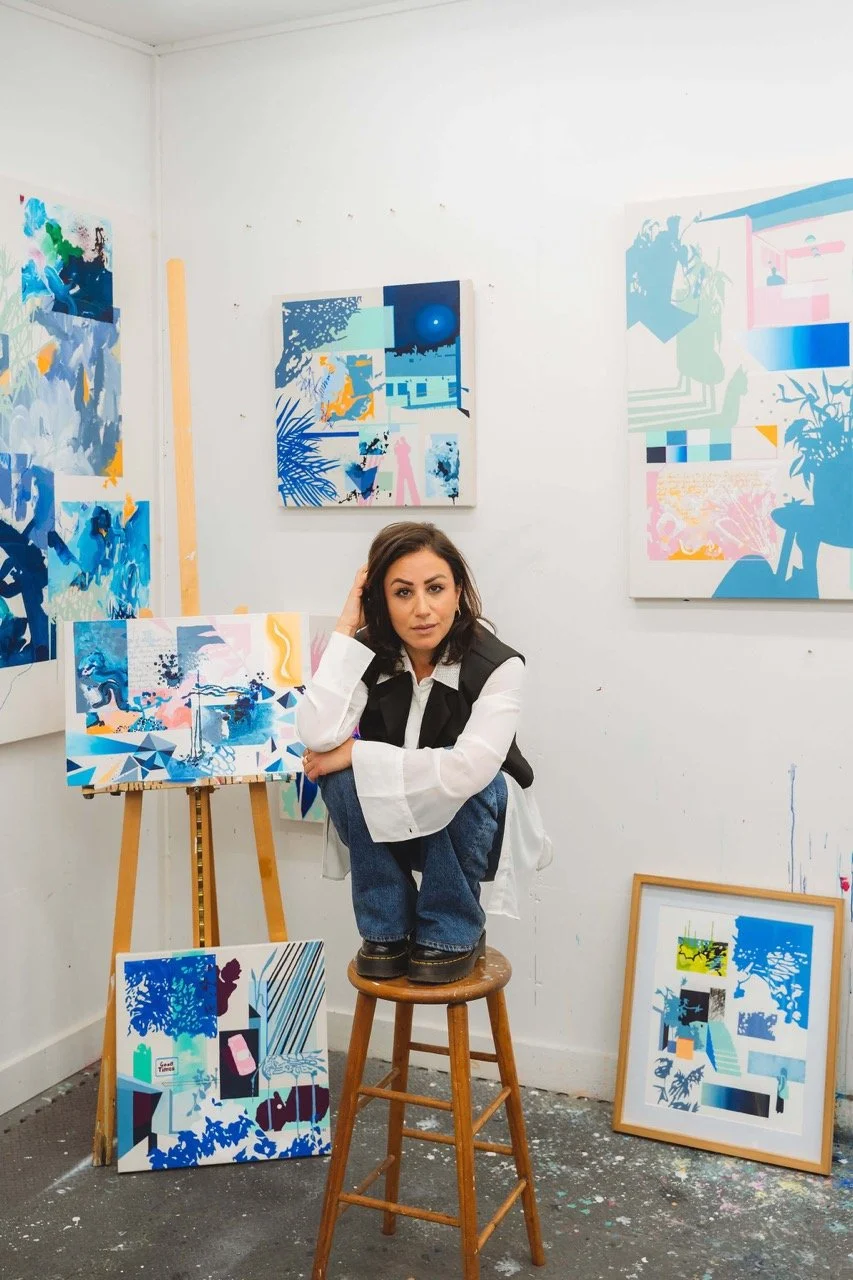 A woman squatting on a wooden stool in an art studio, surrounded by colorful abstract paintings on the walls and floor.