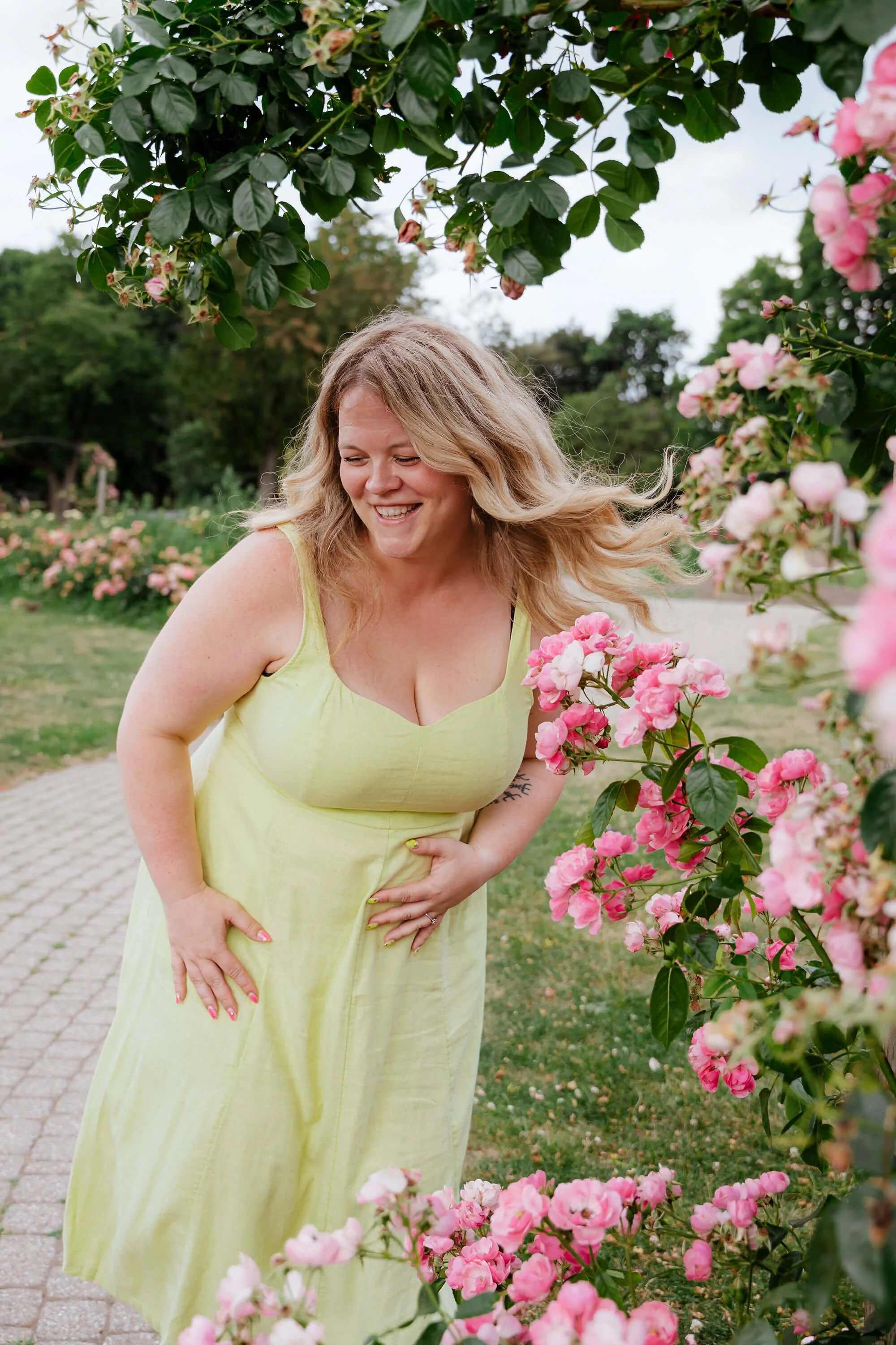 A woman with blonde hair smiling and laughing, wearing a yellow dress, standing outdoors near pink and white flowering bushes.