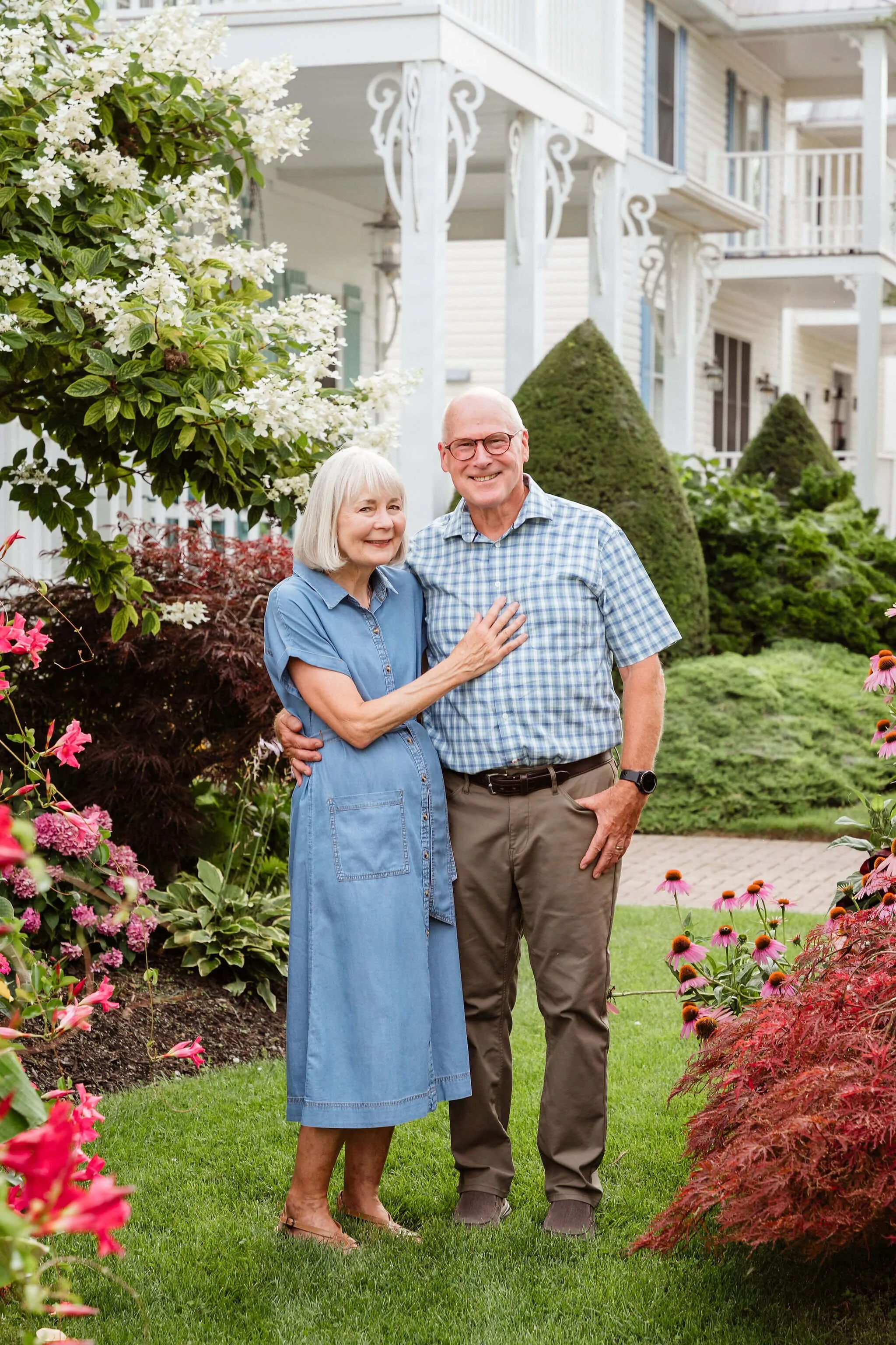 An elderly couple standing in a well-maintained garden with colorful flowers and bushes, smiling at the camera, in front of a white house with a porch and decorative support beams.