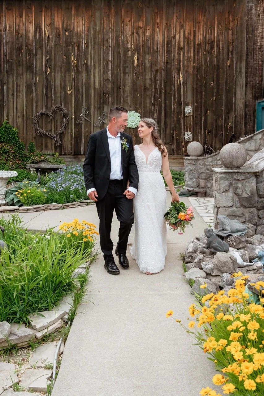 A newlywed couple walking hand in hand on a garden path, with the bride carrying a bouquet of flowers, against a rustic wooden backdrop decorated with heart and sun ornaments.