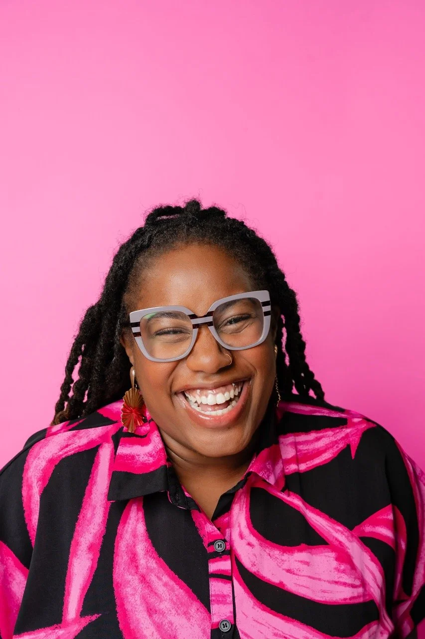 A woman with curly hair, wearing large glasses, colorful earrings, and a vibrant pink and black patterned shirt, smiling joyfully against a pink background.