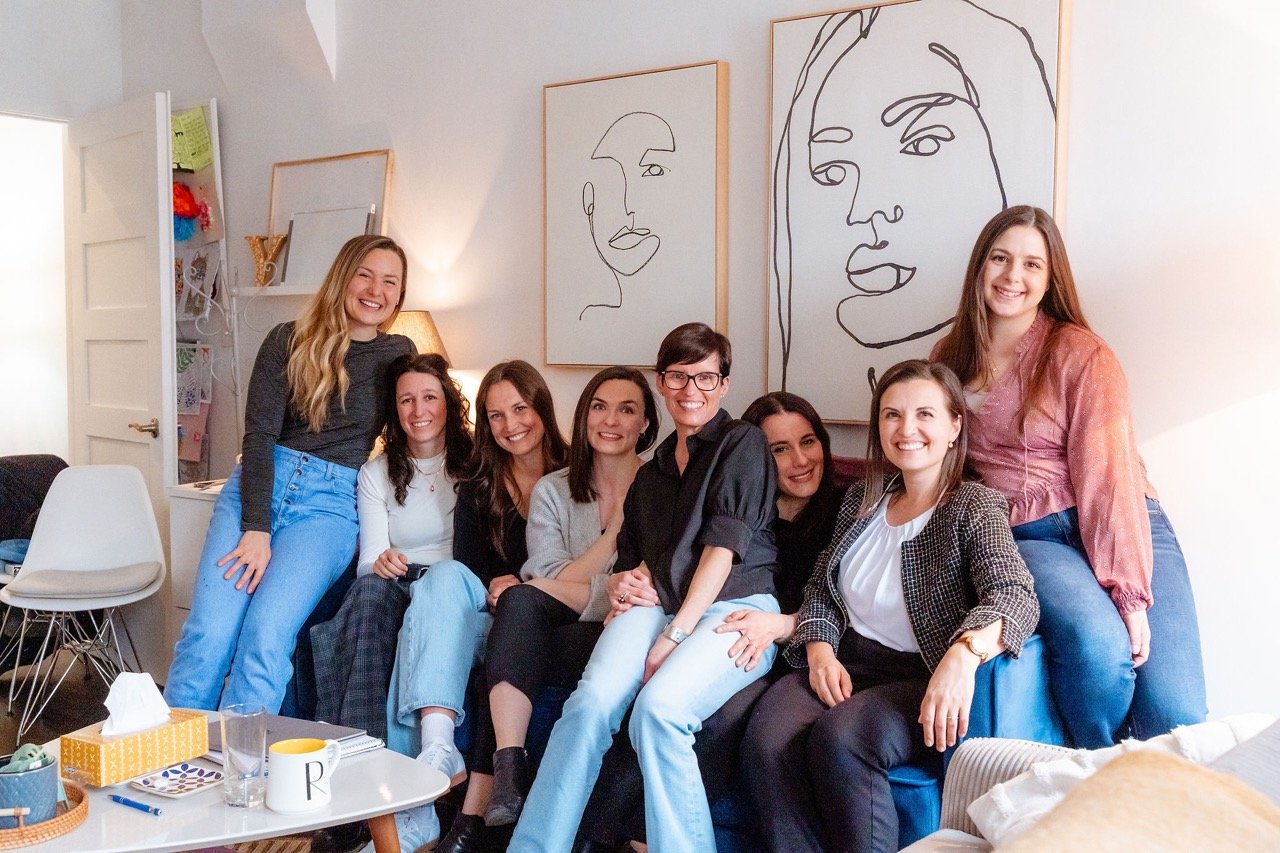 Group of nine women smiling and sitting on a sofa in a cozy, well-lit room with minimalist art on the wall.