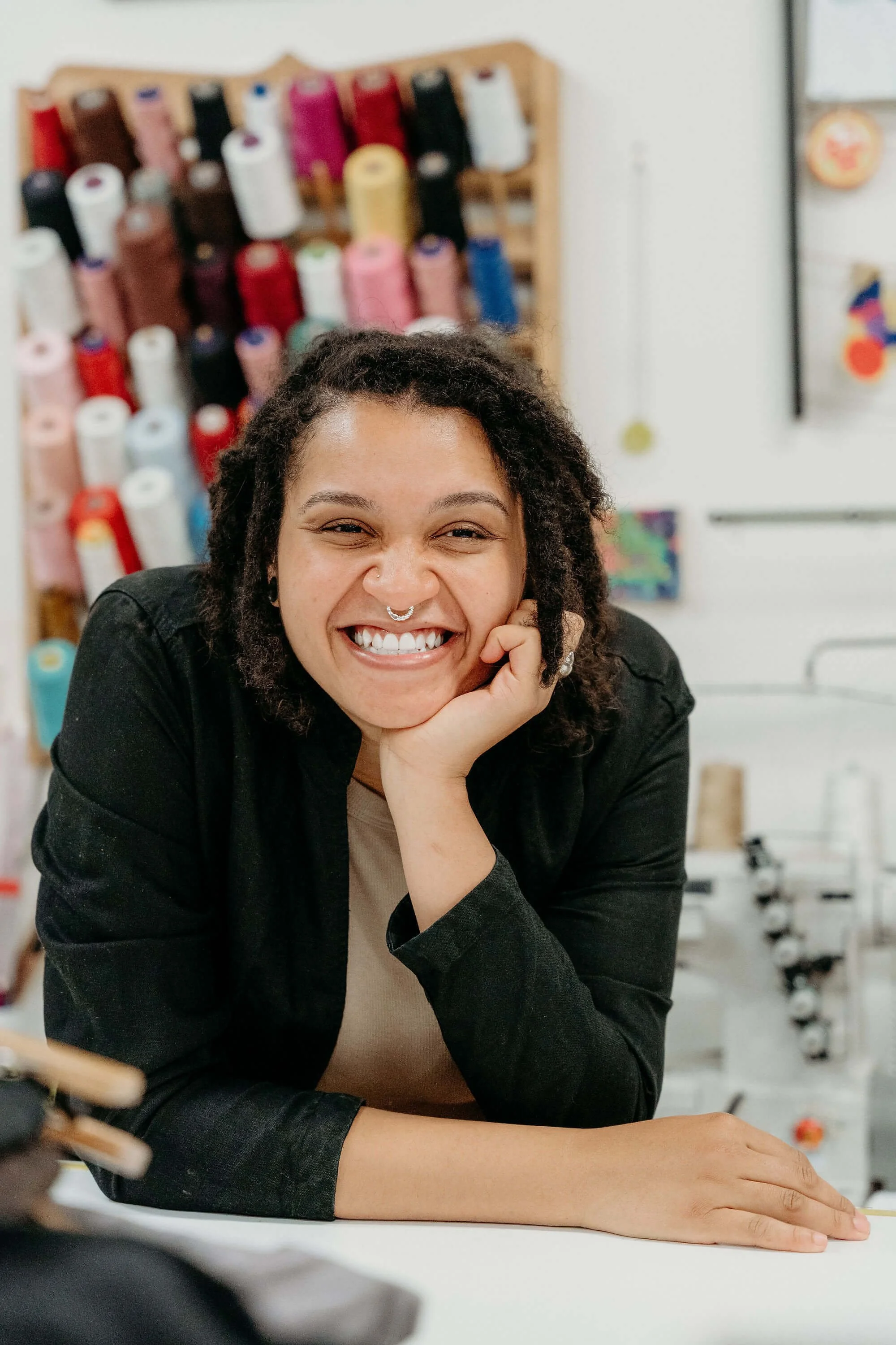 A woman with curly hair smiling, resting her chin on her hand, in a space with colorful thread spools on a rack in the background.