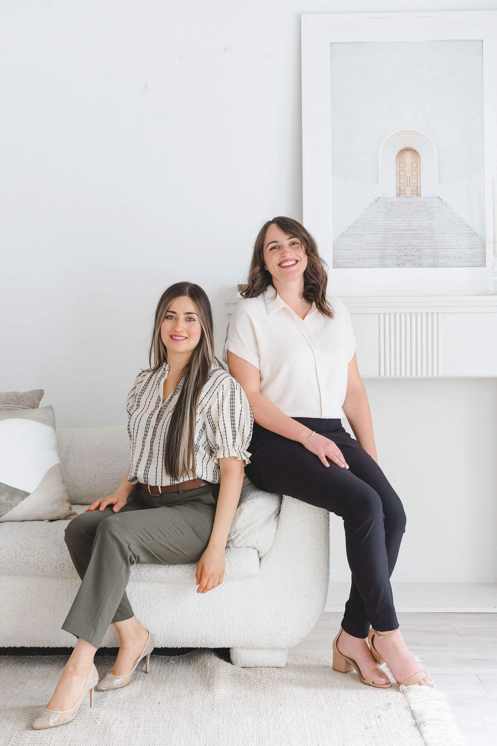 Two women sitting on a white sofa in a bright, minimalist room with a white wall and a large artwork of a staircase and doorway in the background. Both women are smiling, one with long brown hair wearing a striped blouse and gray pants, and the other with curly brown hair wearing a white blouse and black pants.