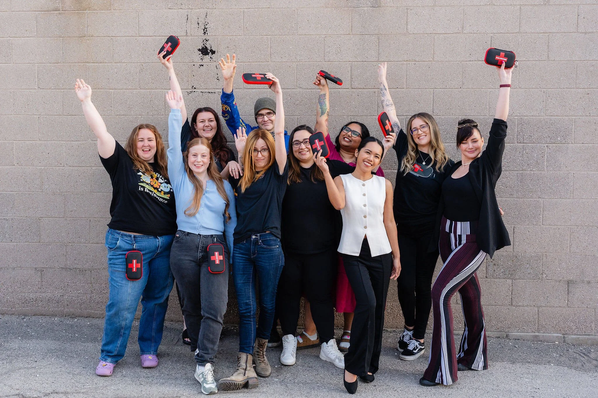 Group of ten diverse young women standing outdoors against a concrete wall, smiling and celebrating with raised arms, some holding Red Cross first aid kits.