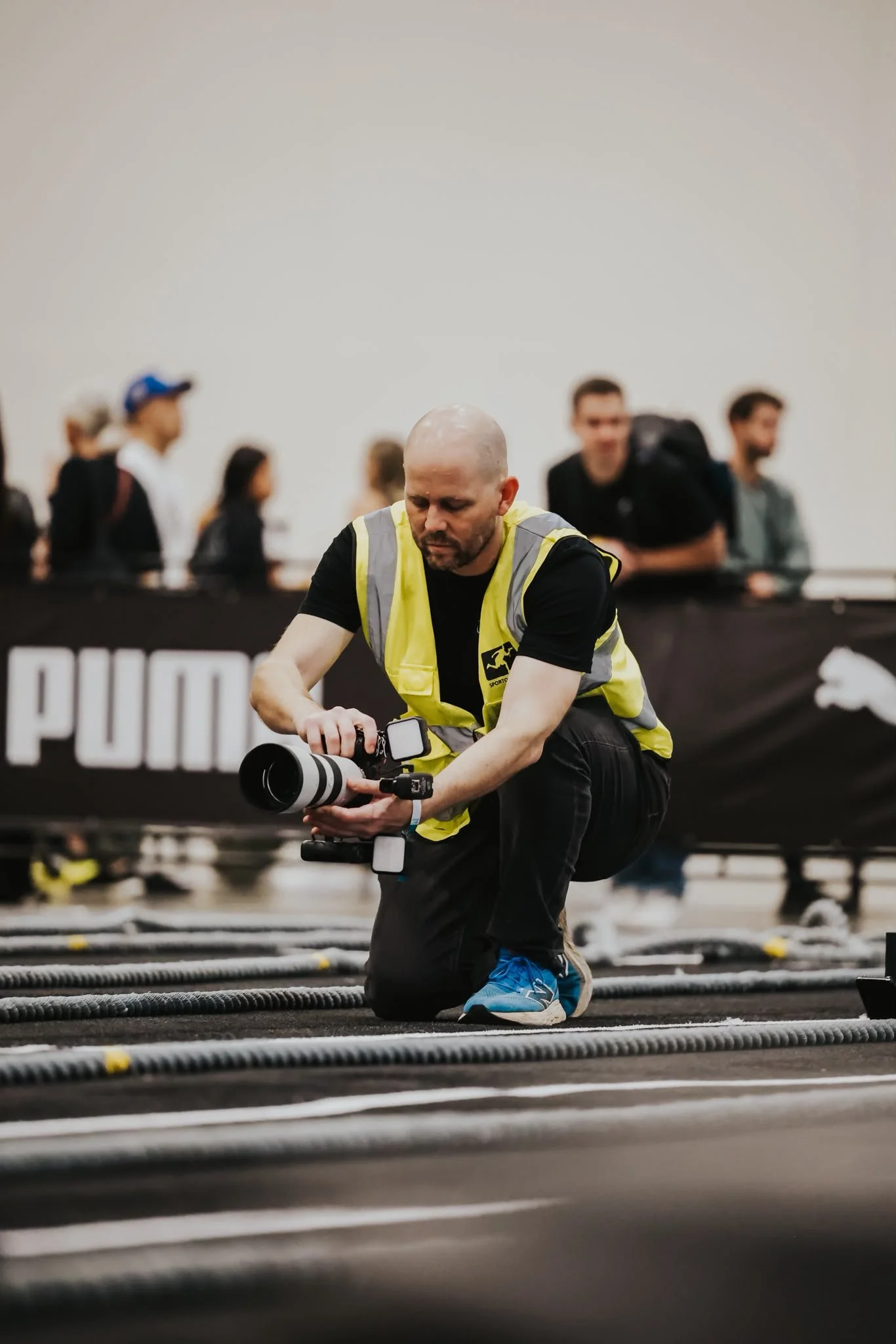 A photographer kneels down at a hyrox even between ropes laying on the floor, holding a camera for a low angle shot of an athlete.