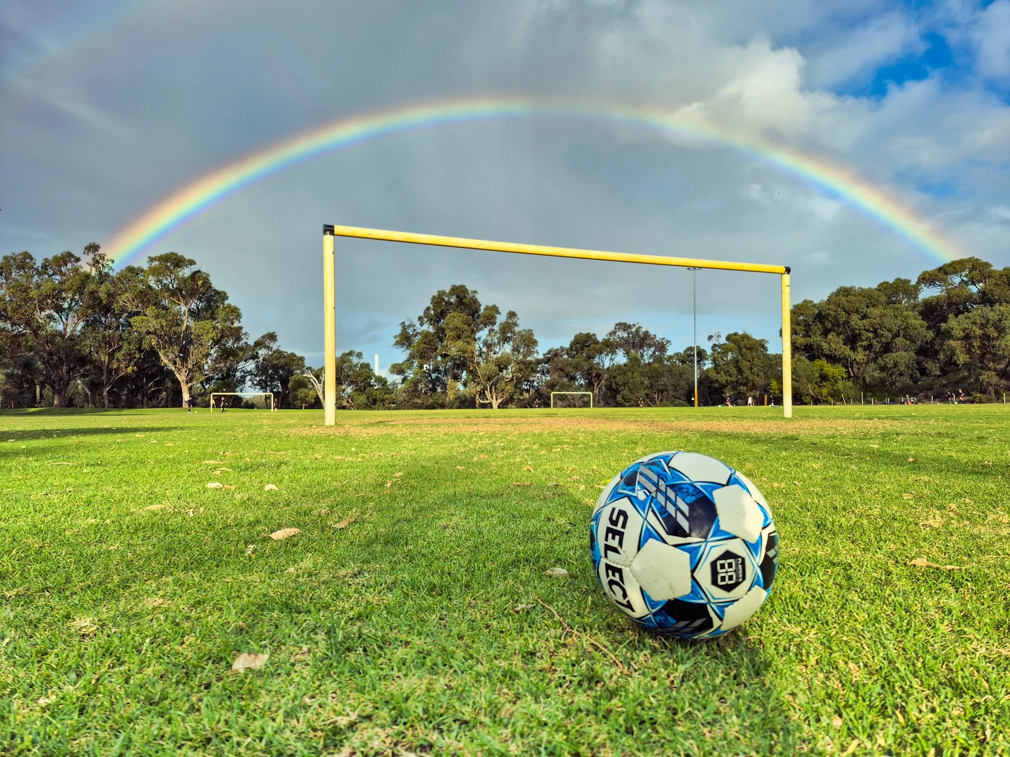 A soccerball sits on a football field in front of a set of soccer goalposts while a rainbow shines overhead