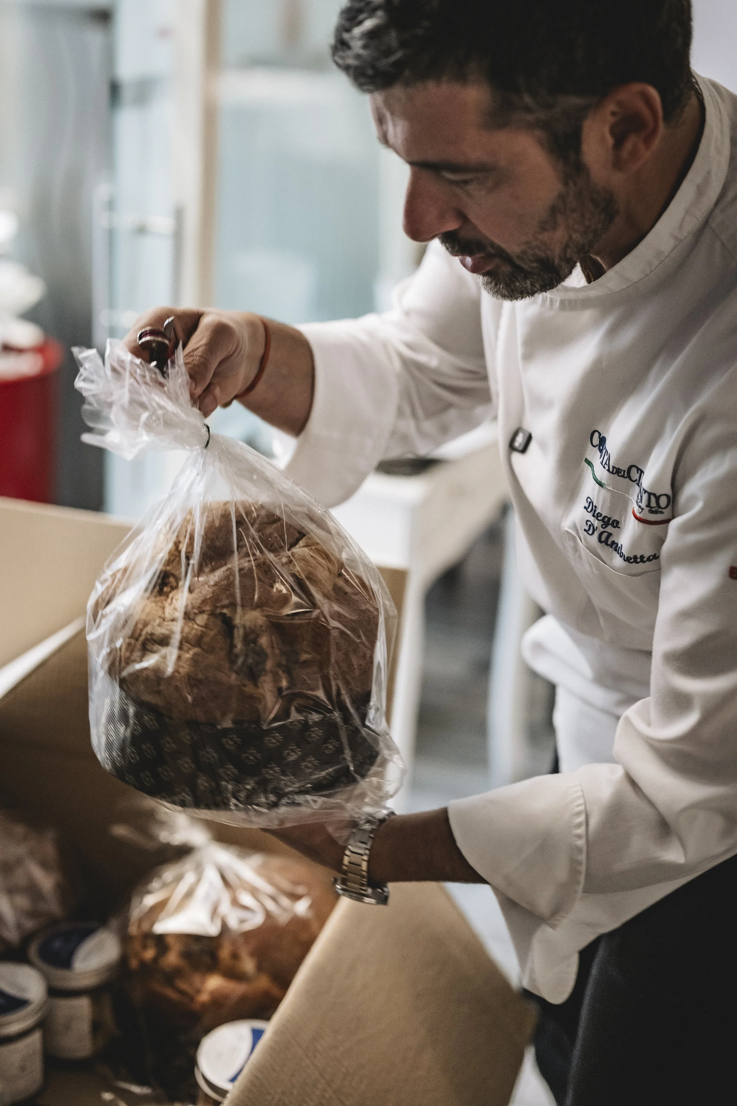 Uno chef con giacca bianca e orologio al polso apre una busta trasparente con un pane o una focaccia dentro, in un ambiente di cucina o laboratorio alimentare.