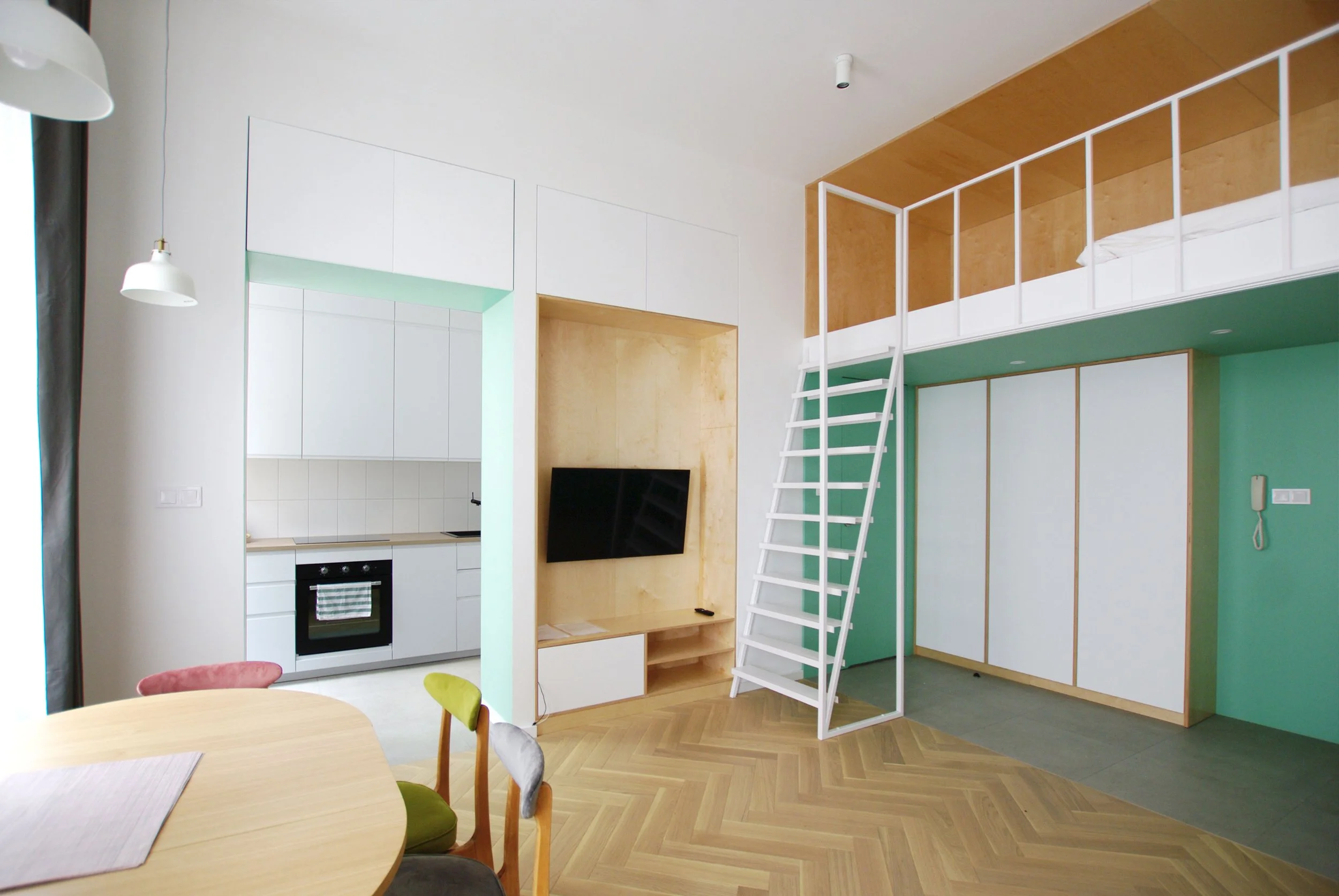 Open-plan interior at Apartment 30, with a white kitchen, timber dining table, herringbone flooring and a mezzanine sleeping area.