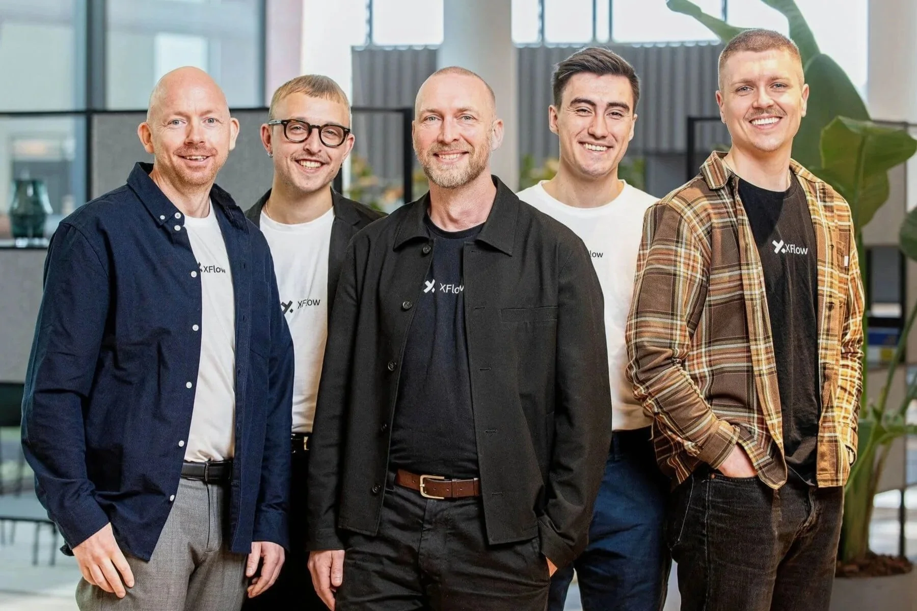 Fem mænd står sammen i et kontormiljø, alle smilende og iført afslappet tøj med logoet XFlow. Baggrund med store vinduer og grønne planter.