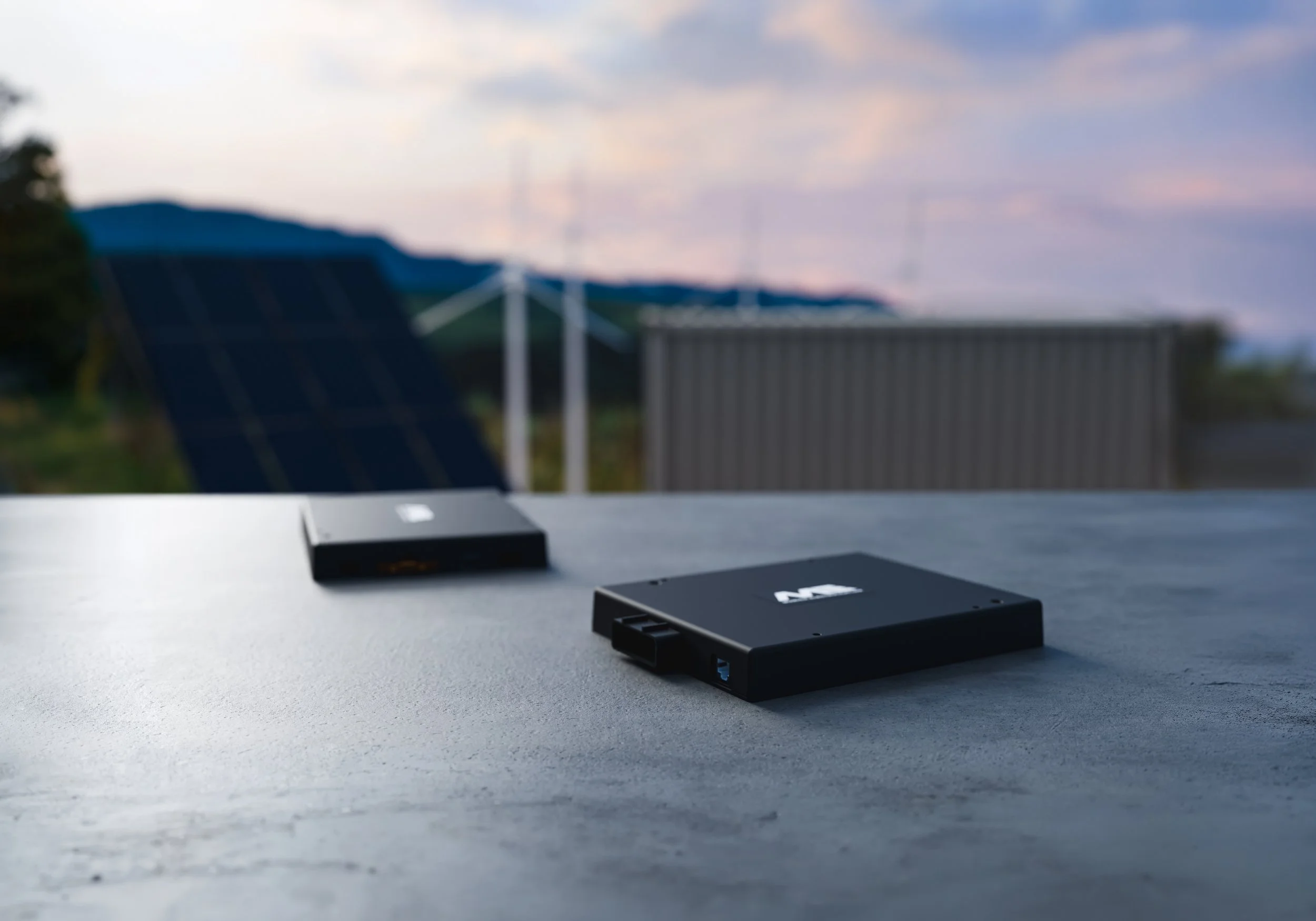 Two Stationary ESS Battery Management Systems laying on a concrete surface, the background shows a solar panel, wind turbines and a BESS container