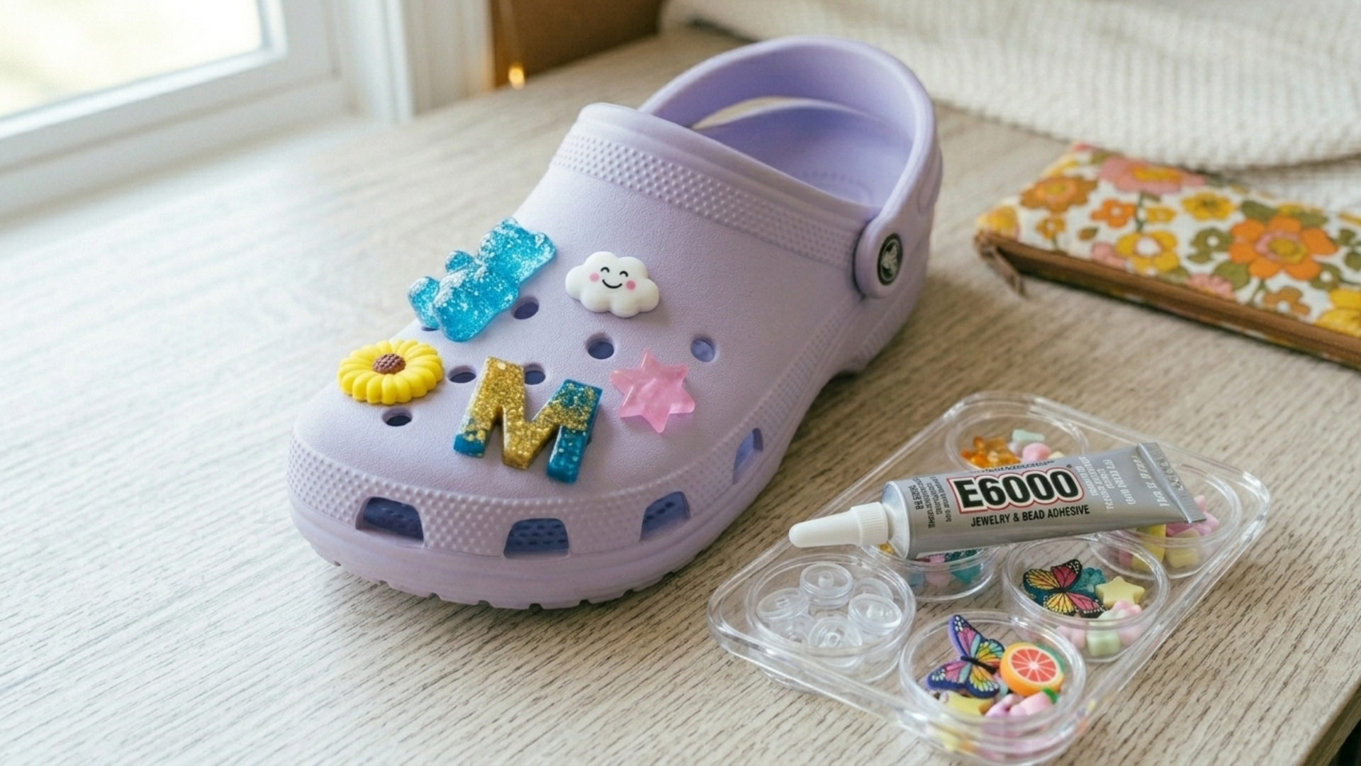 A charm bar party setup with a Croc-style clog in purple adorned with DIY Jibbitz. A clear muffin tin with different beads and charms sits next to the shoe on the table, along with a tube of E6000 glue.