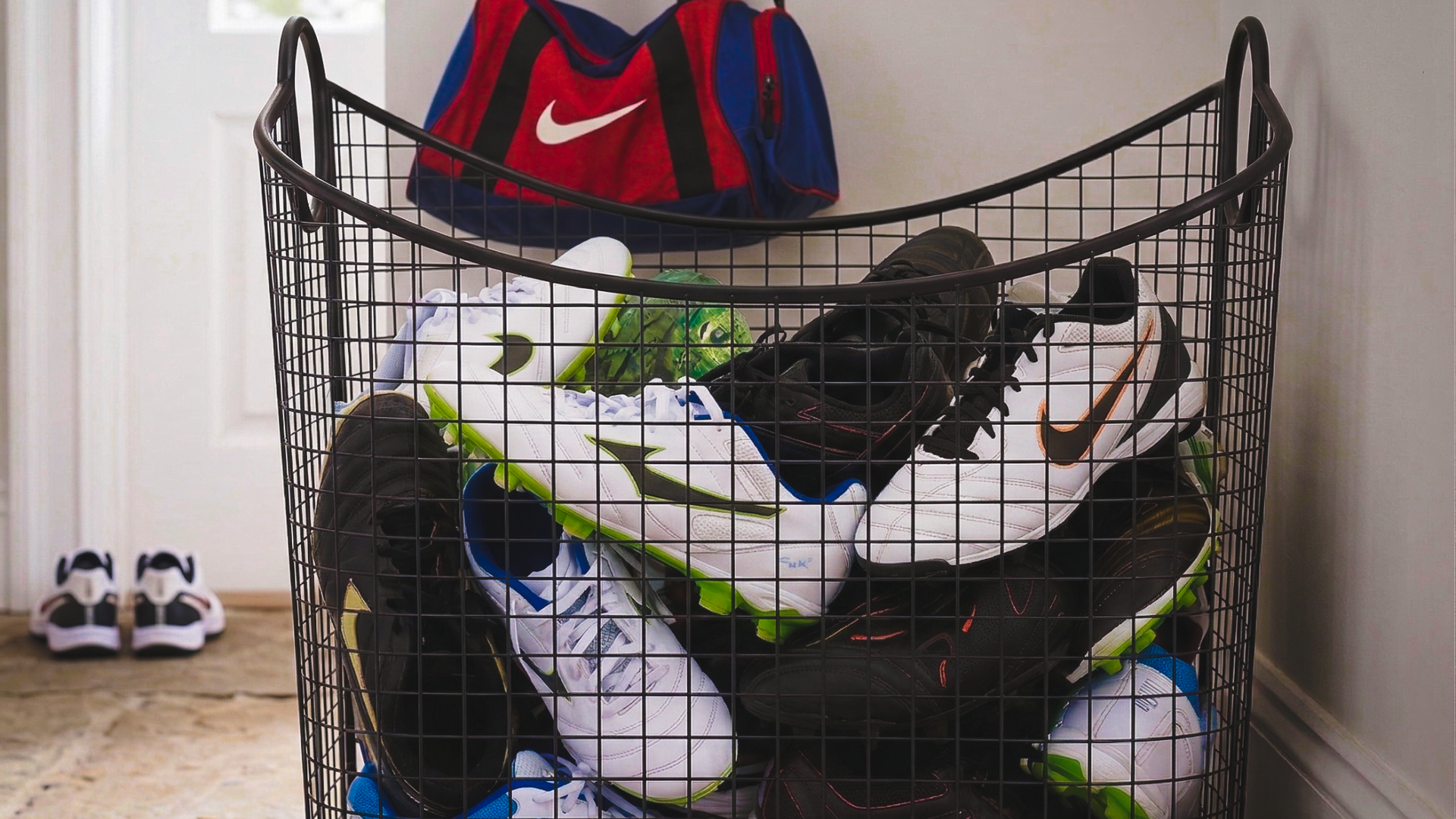 A close-up view of a home entryway. A black mesh basket with baseball, softball, and soccer cleats tossed inside for convenient storage. A sports duffel bag hangs on the wall, showing different ways to store and organize kids' sports gear.