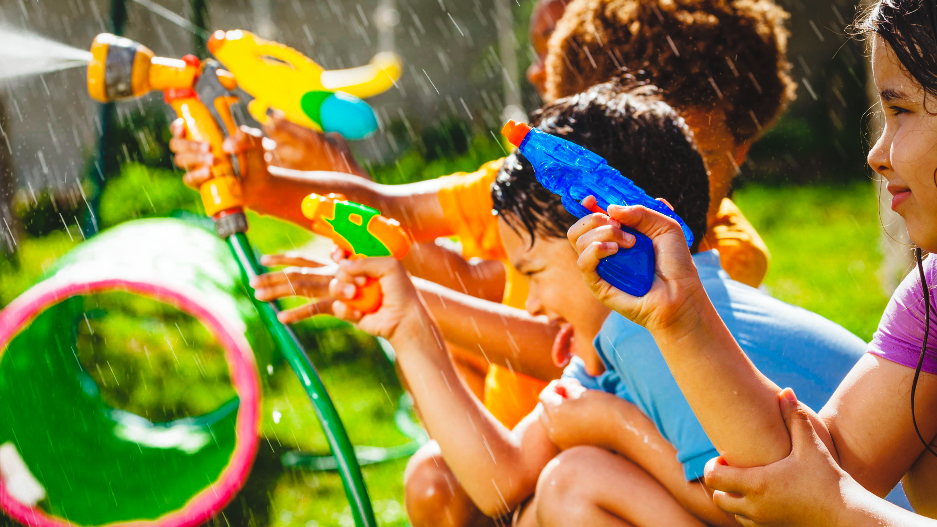 A group of children in a sunny backyard playing with colorful water guns and sprayers near a green play tunnel.