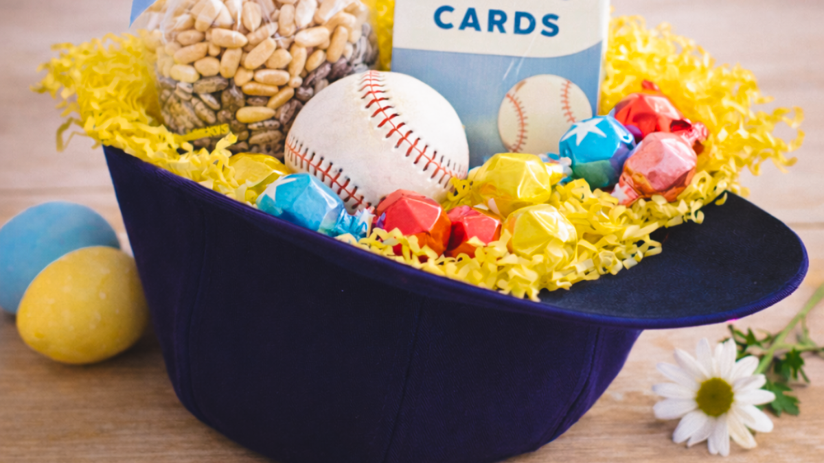 A navy blue baseball hat used as a creative Easter basket, filled with yellow crinkle paper, a baseball, trading cards, and wrapped candies, sitting on a wooden surface with a daisy and Easter eggs.