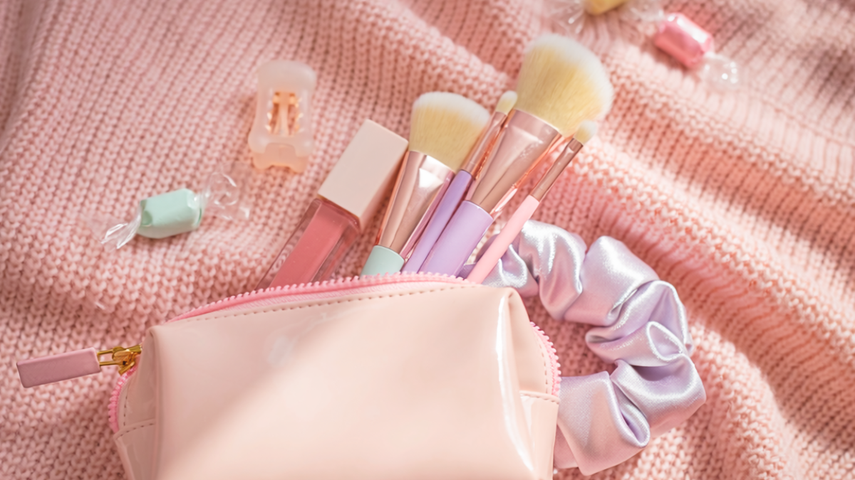A light pink glossy makeup bag used as a creative Easter basket alternative, filled with makeup brushes, lip gloss, a satin scrunchie, and hair clips, styled on a pink knit sweater.