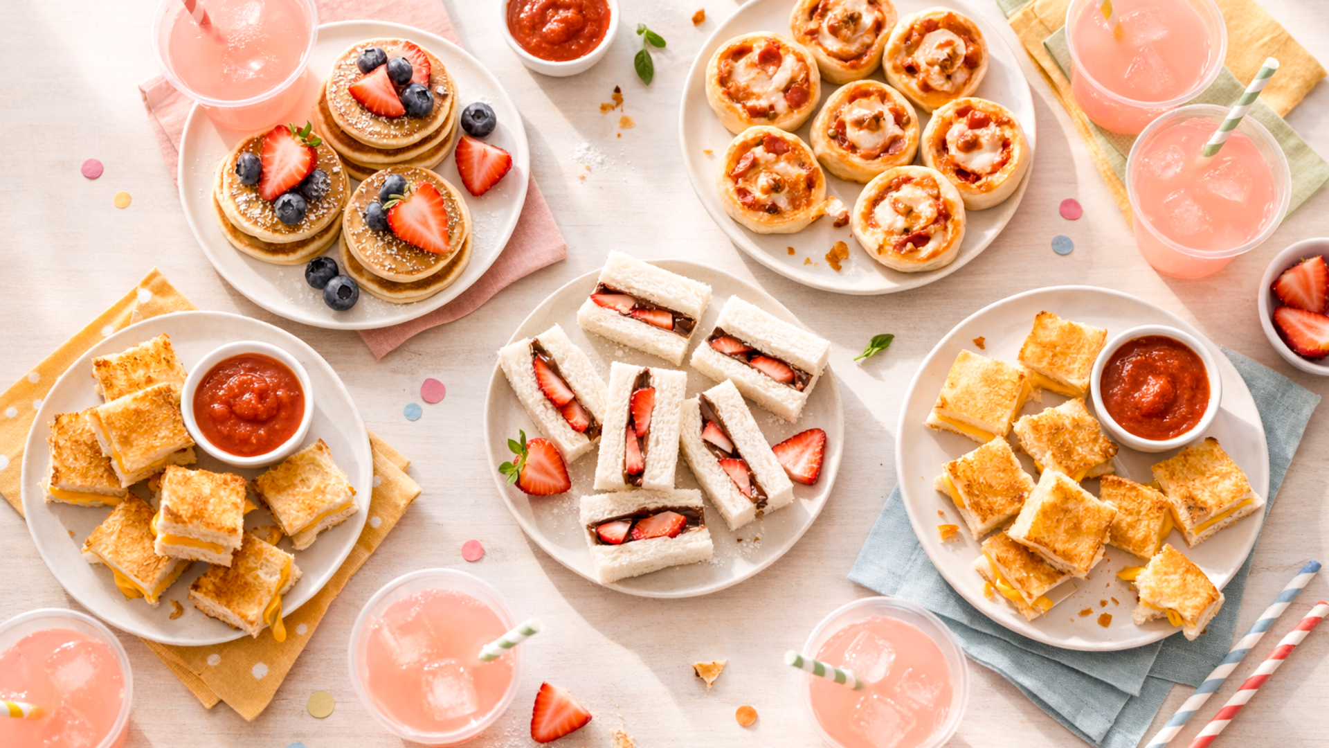 Kids' tea party food setup with easy snacks and colorful drinks. Overhead flat lay shot shows strawberry and nutella sandwiches, mini pancakes, and grilled cheese bites.