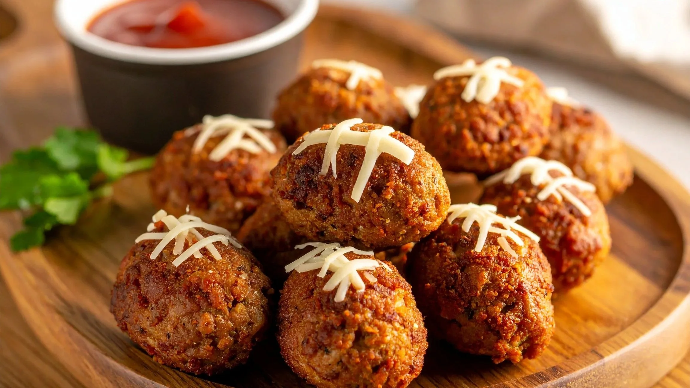 A tray of meatballs topped with cheese laces sits on a wooden serving tray next to a bowl of dipping sauce, ready to be eaten by a Super Bowl party crowd
