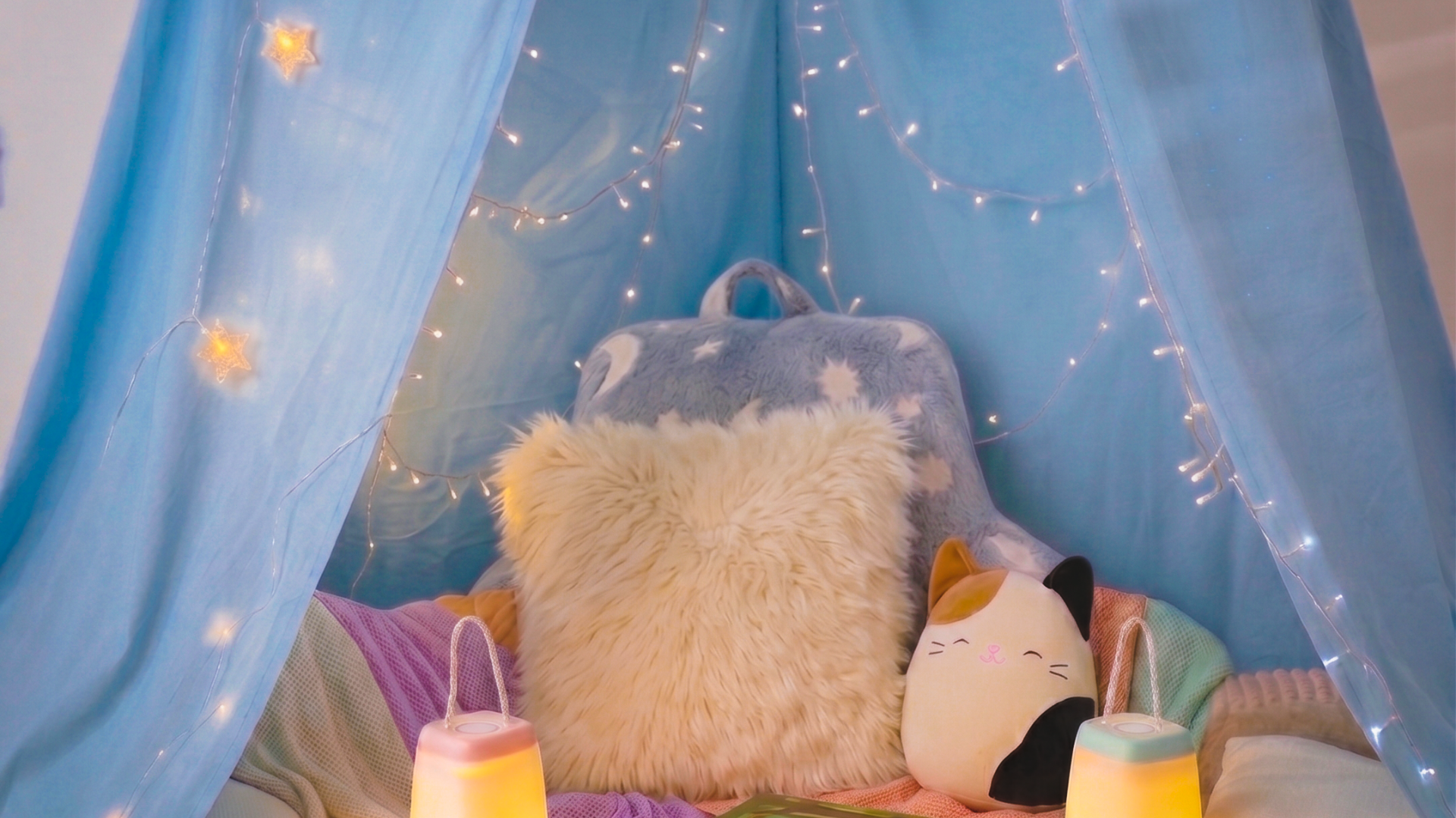 Child's reading nook setup with cozy pillows and blankets, a Squishmallow stuffed animal, soft lights, and a stack of reading books, all nestled in a soft blue indoor tent with fairy lights.